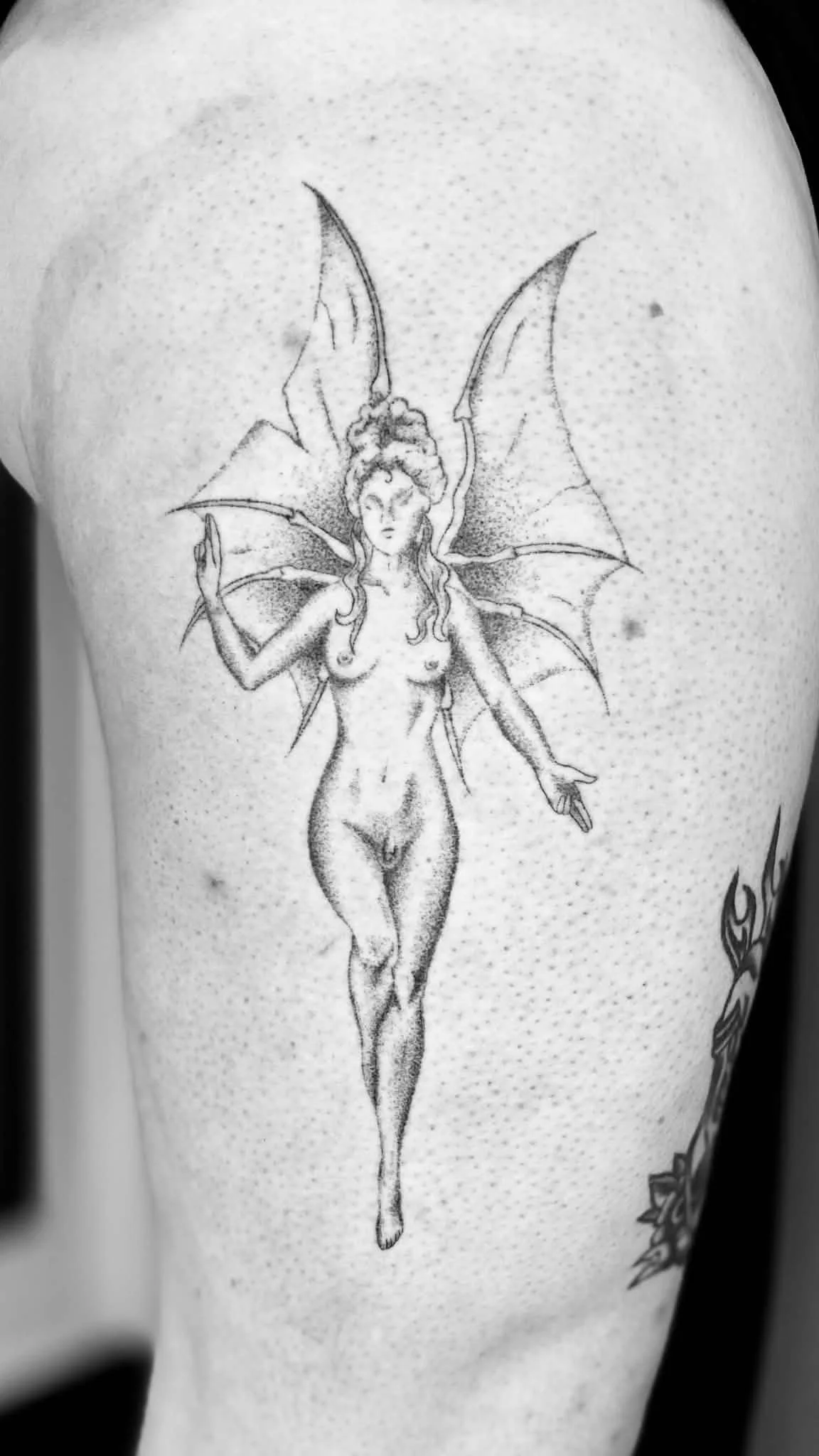 Black and white tattoo of a fairy with bat-like wings, standing nude with one arm raised and the other extended downward, on person's arm.