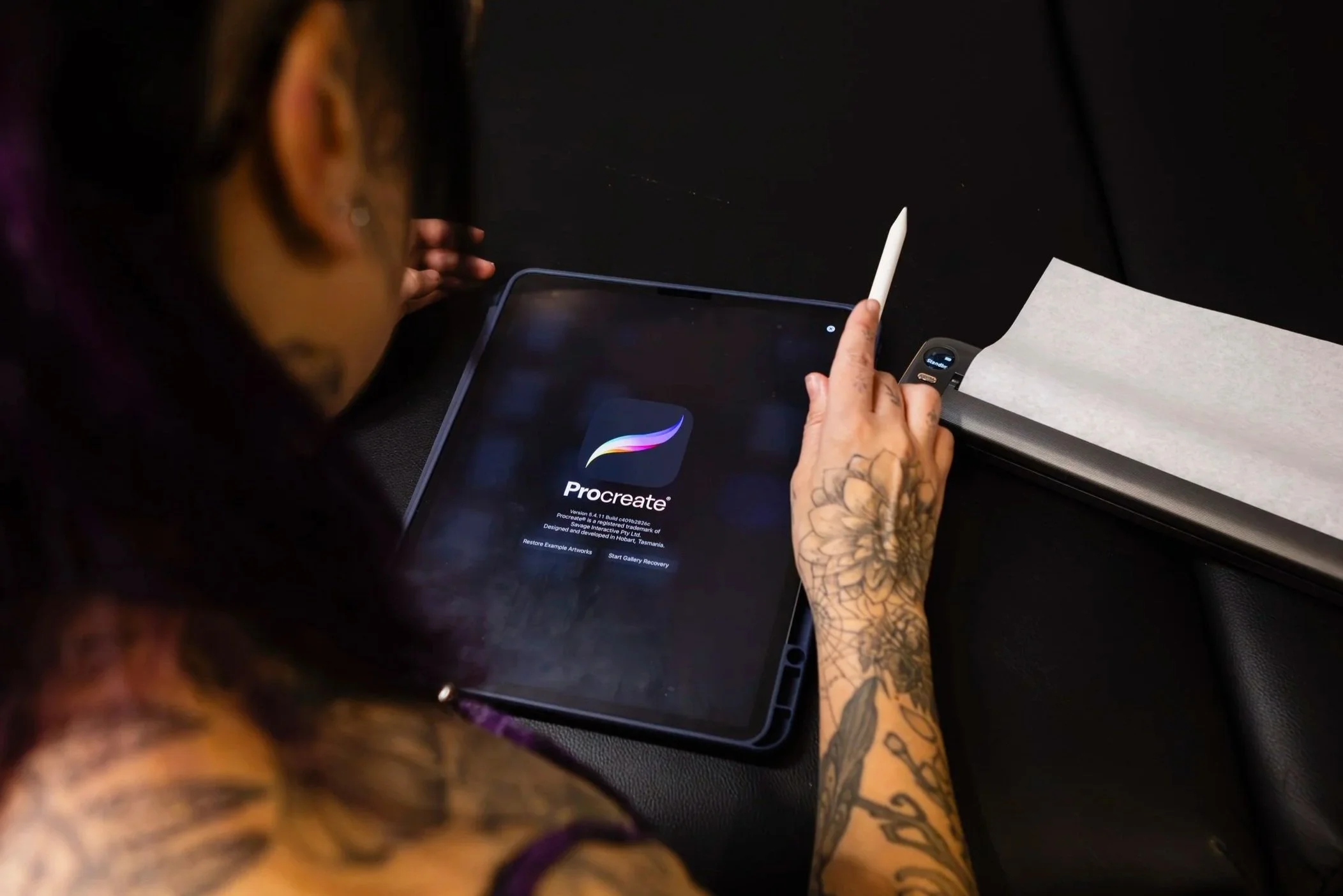 A person with tattoos on their arms using a white stylus and a tablet displaying the Procreate app, sitting at a black table with a sheet of paper and a scanner nearby.
