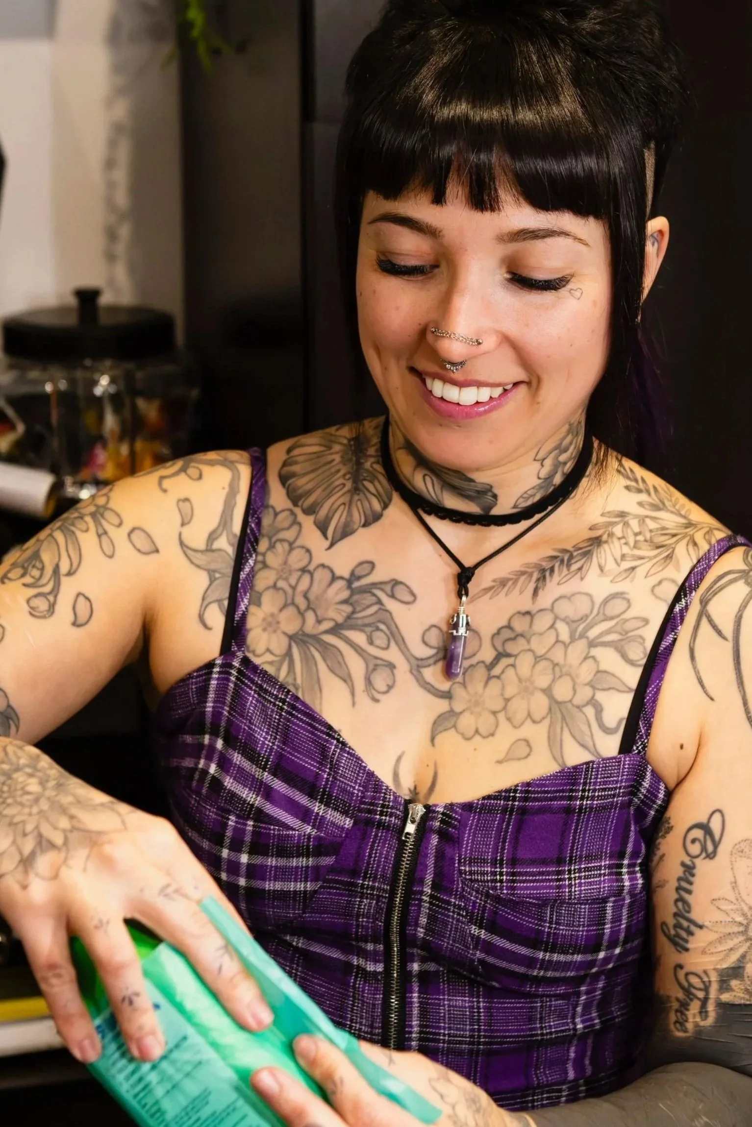 A woman with short black hair, tattoos, and piercings smiling while holding a green item in her hand. She is wearing a purple plaid top and multiple necklaces.