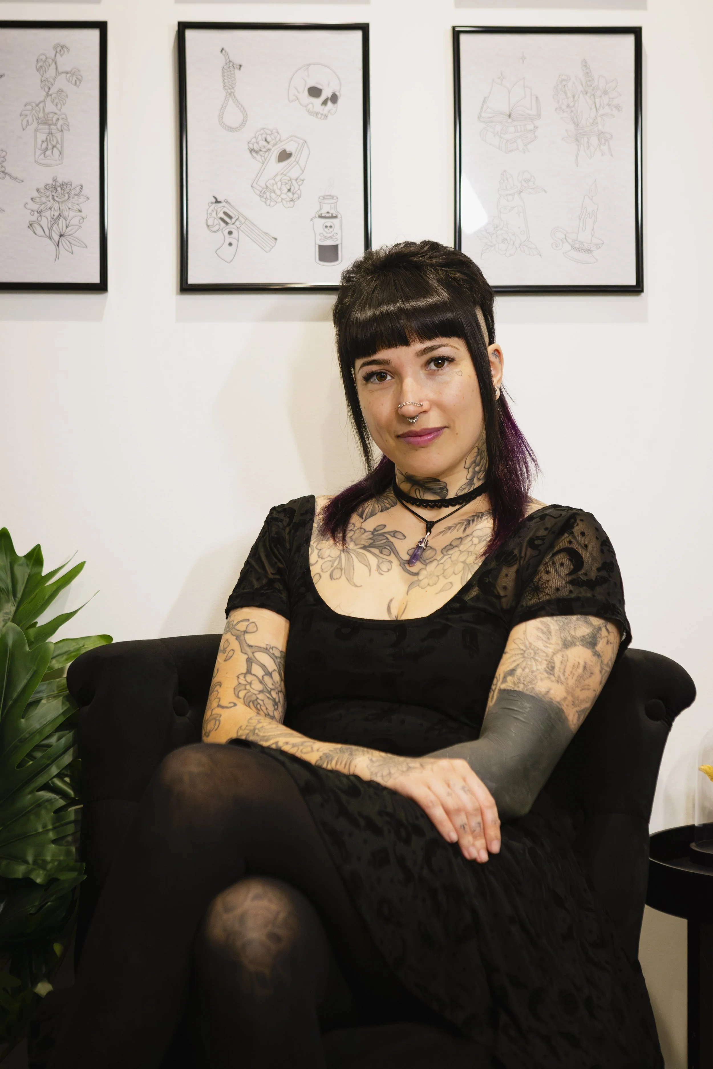A woman with dark hair and tattoos sitting on a black velvet chair in an art gallery, with framed tattoo-inspired artwork on the wall behind her, and a green plant to her side.