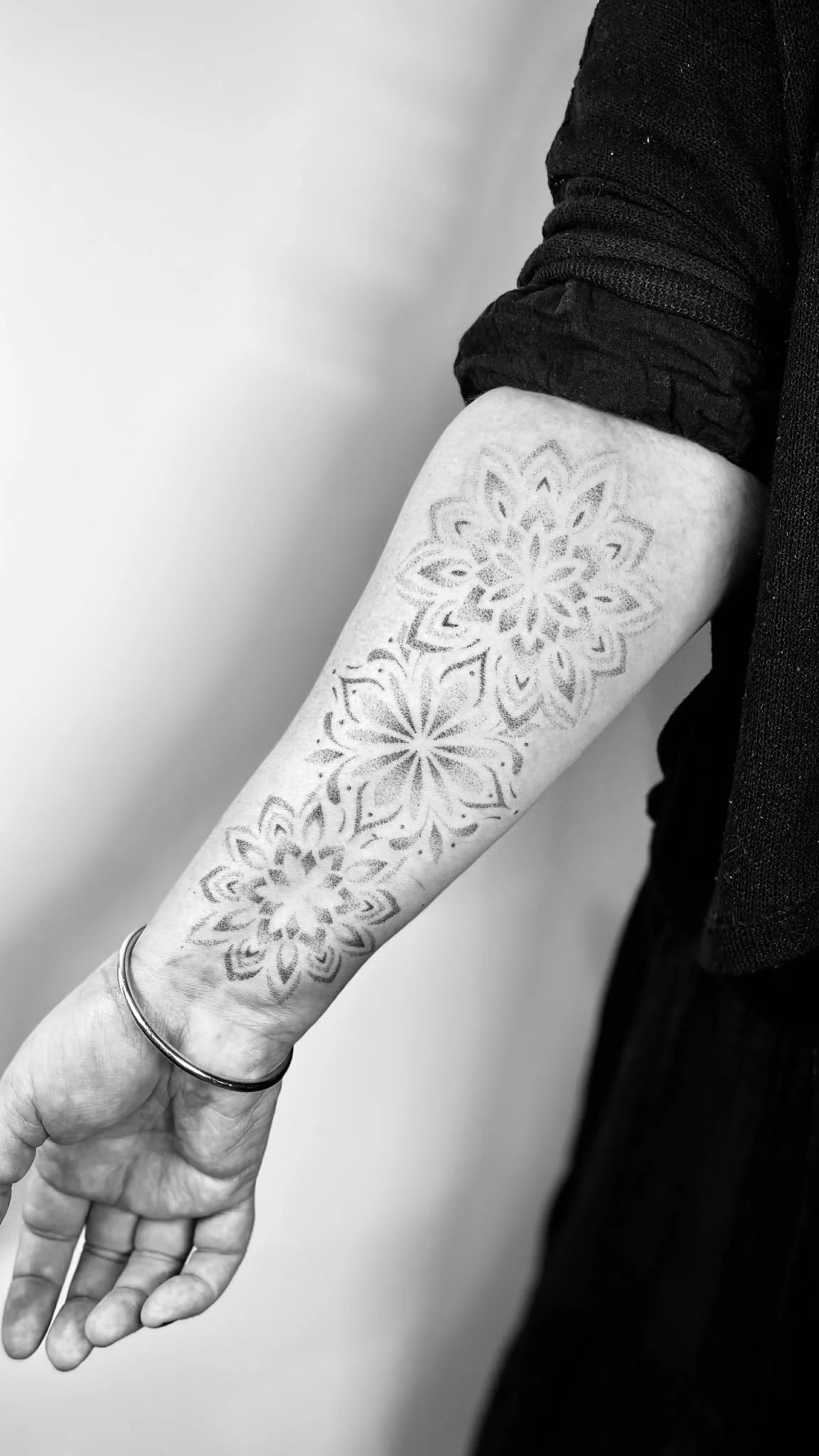A person's arm with a floral tattoo in black and white, wearing a black long sleeve shirt with the sleeve rolled up, a bracelet, and a ring.