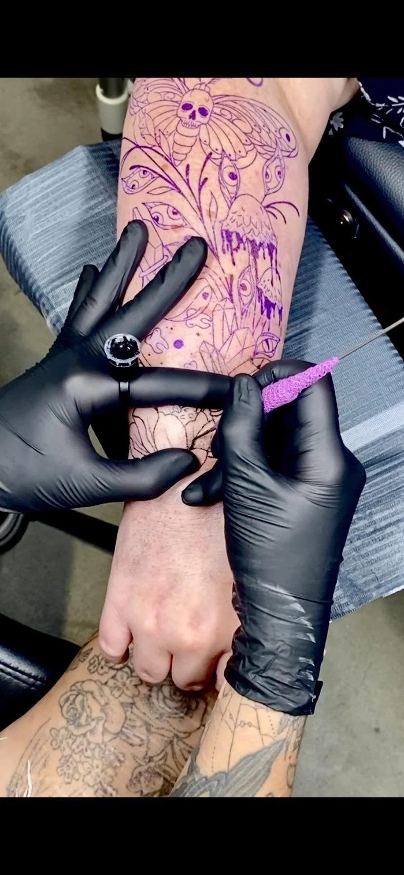 A person is getting a tattoo on their leg, with the tattoo design. The tattoo artist, wearing black gloves and a ring, is holding a needle bar.
