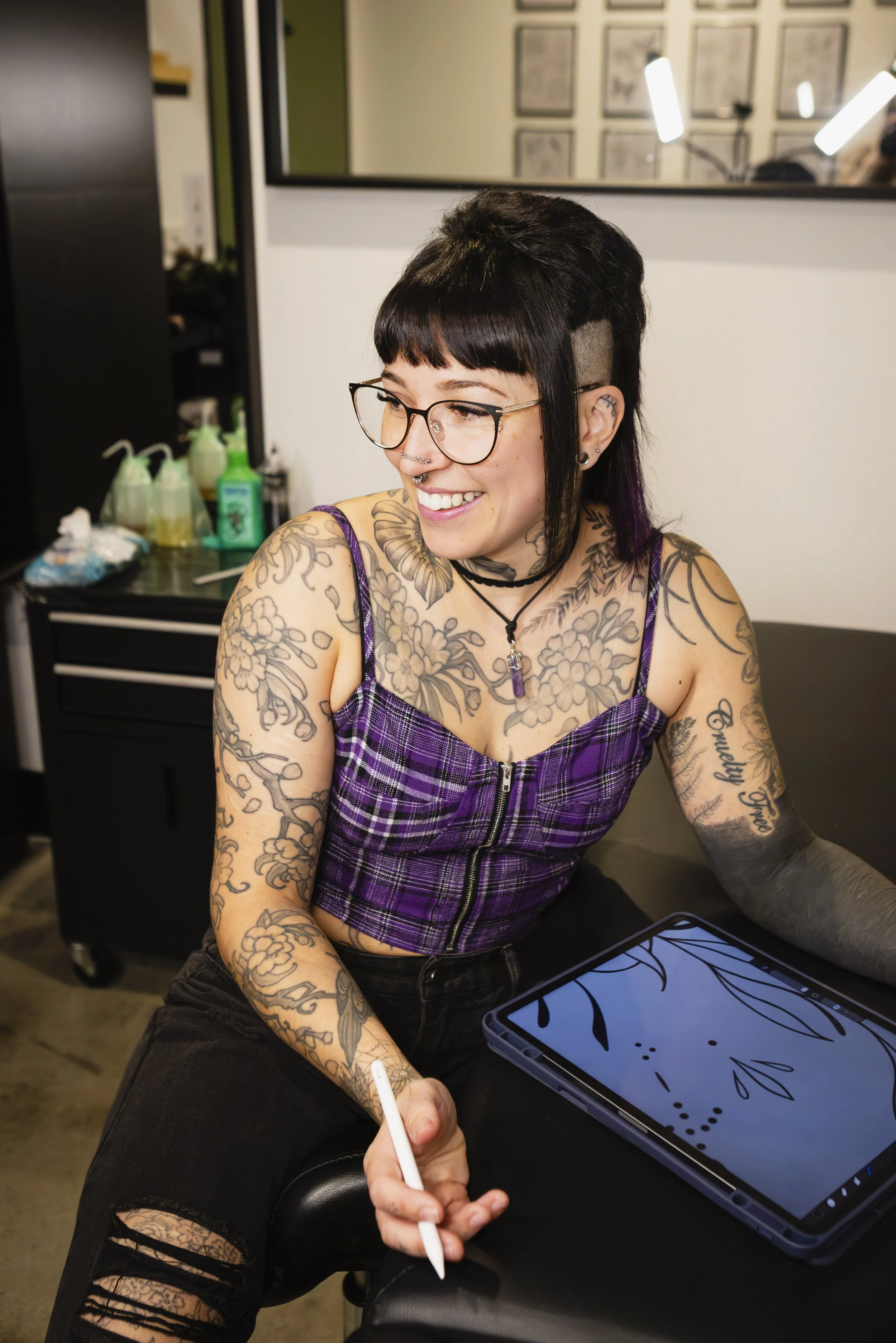 A smiling woman with tattoos on her arms and chest, wearing glasses and a purple plaid top, drawing on a digital tablet with a stylus in a tattoo studio.