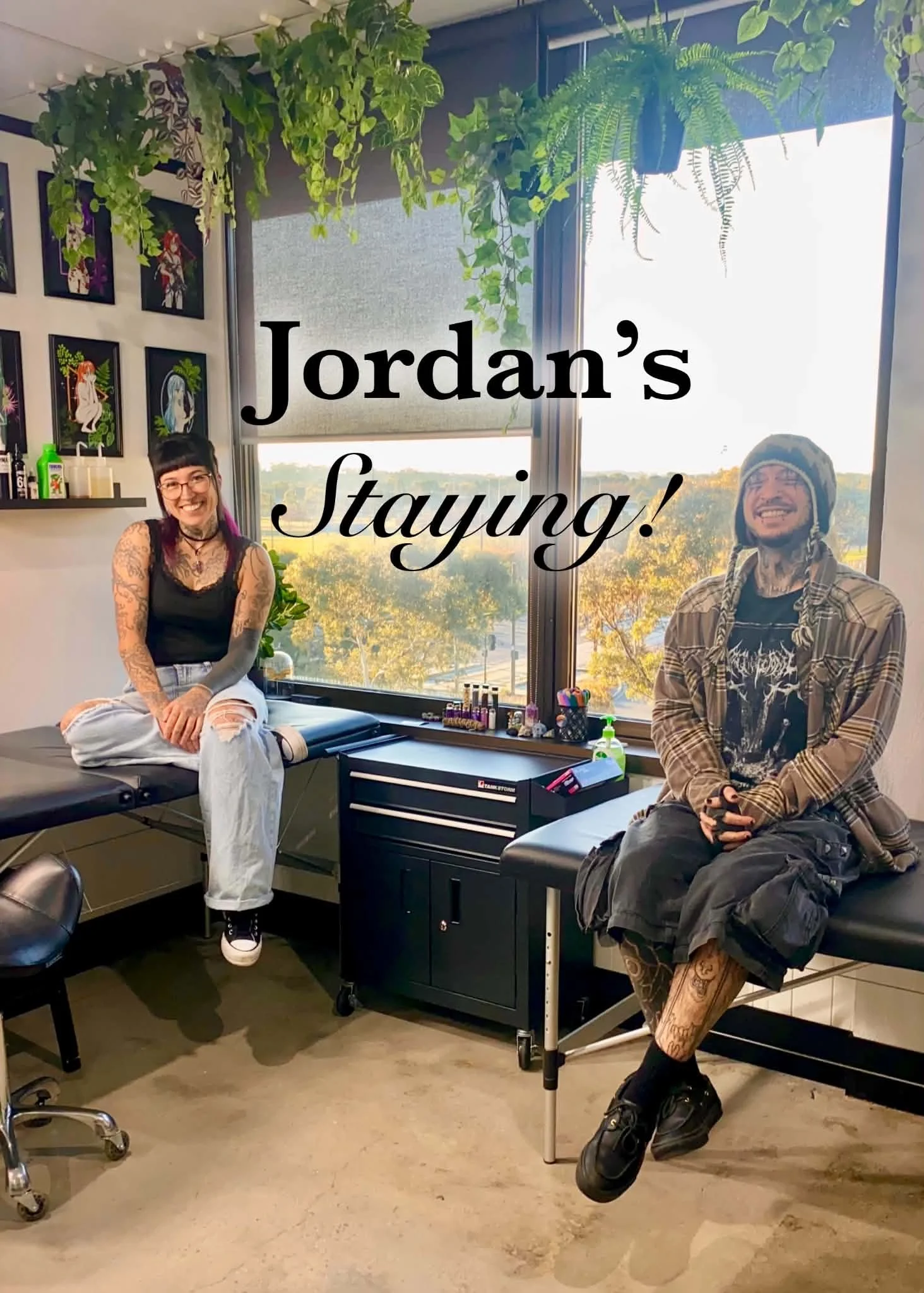 Two people in a tattoo studio, sitting on tattoo chairs. One woman with glasses, black tank top, and tattoos on her arms, smiling. One man wearing a patterned hat, plaid shirt, and shorts, also smiling. The studio has large windows with a view of trees, hanging plants, tattoo artwork on the wall, and tattoo supplies on a counter.
