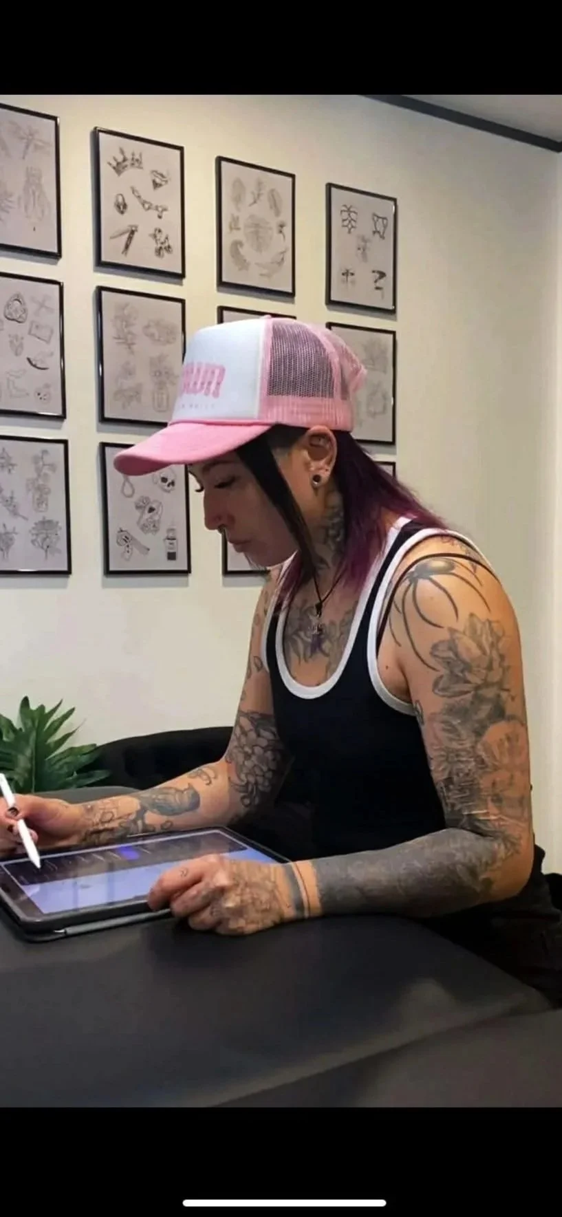 A tattooed woman wearing a black tank top and pink and white cap sitting at a table with a tablet, drawing or writing with a stylus inside a room with framed artwork on the wall behind her.