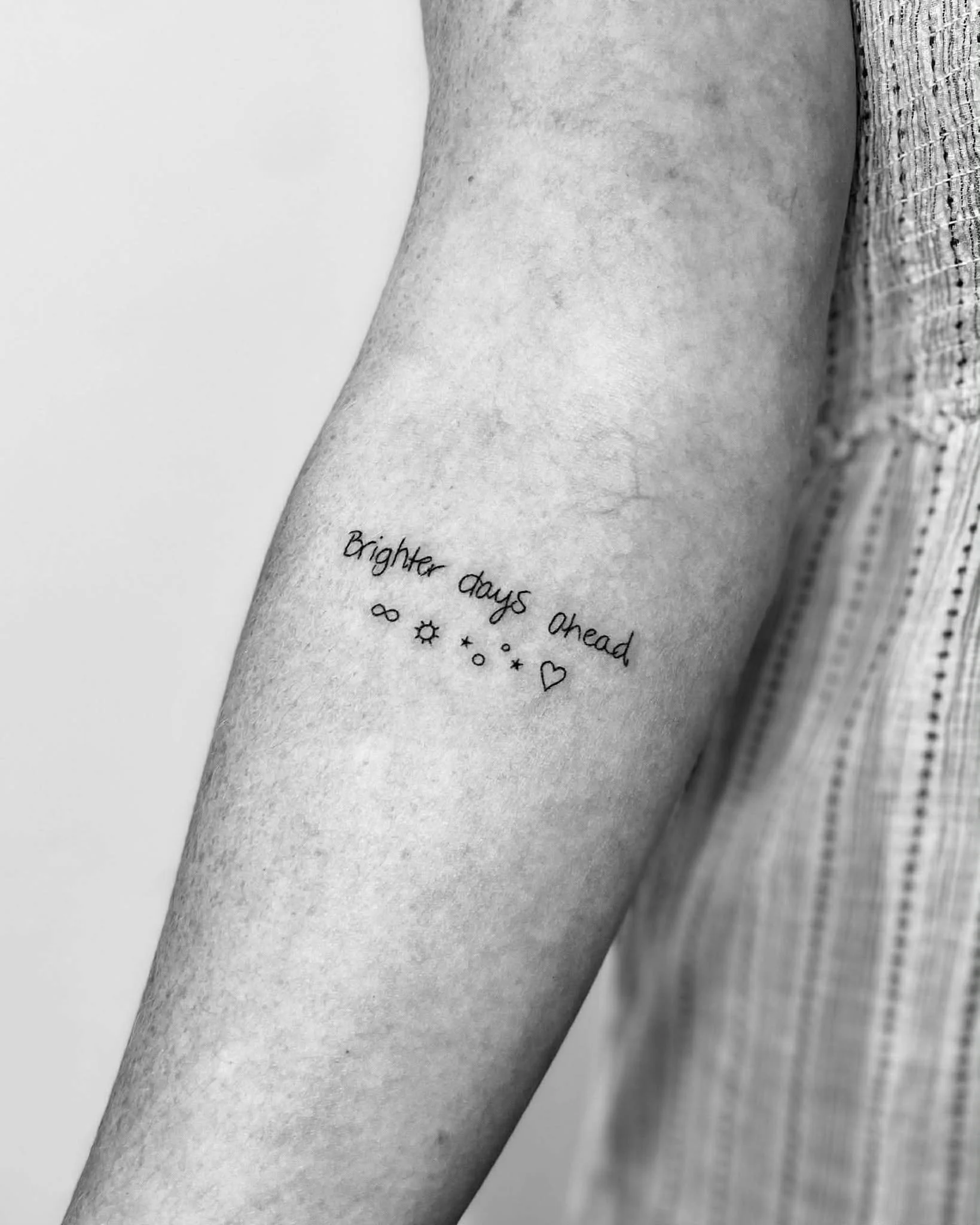 A tattoo on a person's arm that says 'Brighter days ahead' with small decorative symbols including a sun, moon, and hearts.
