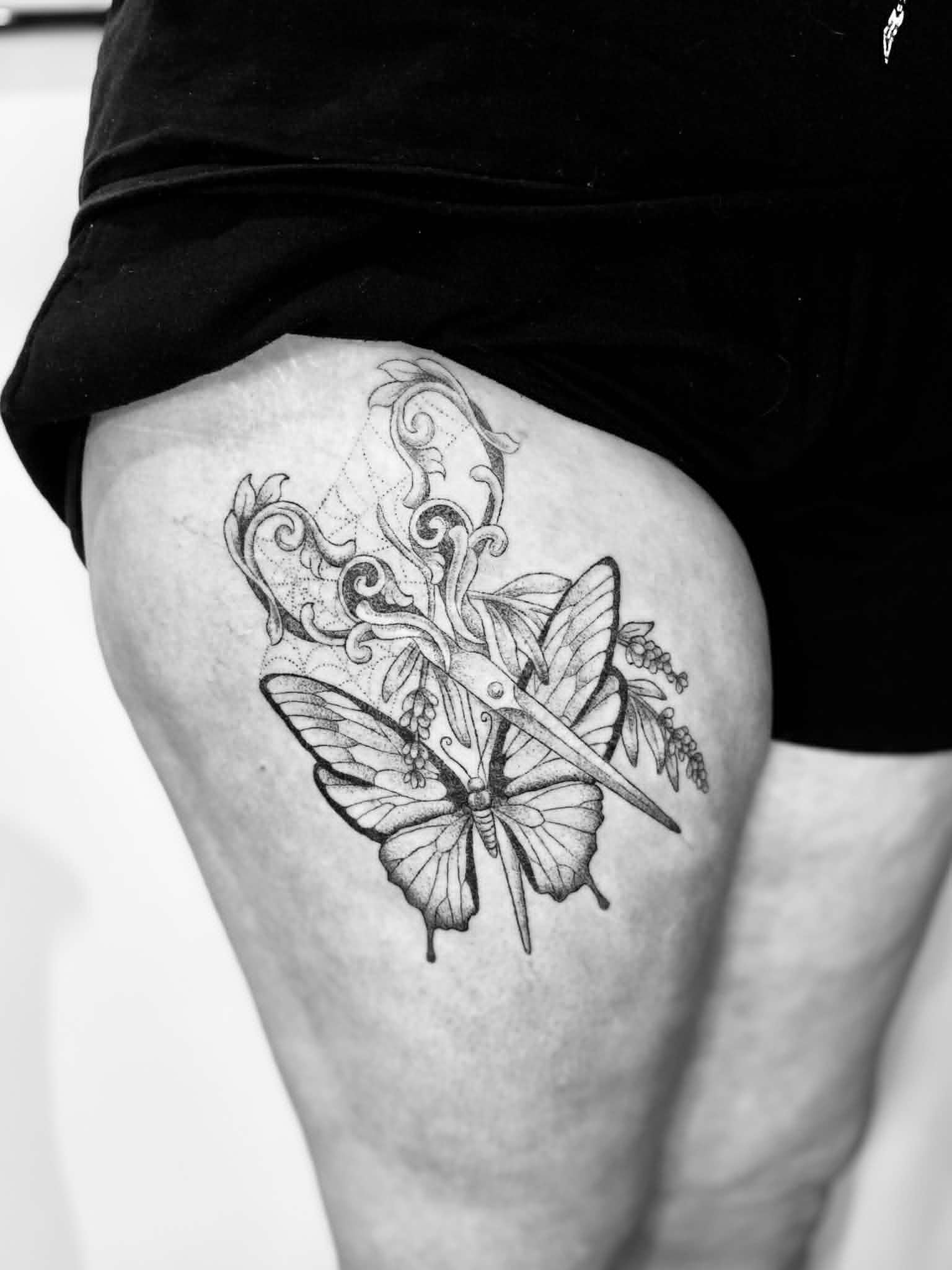 Black and white photograph of a tattoo on a person's thigh, featuring a butterfly with detailed wings and floral elements.