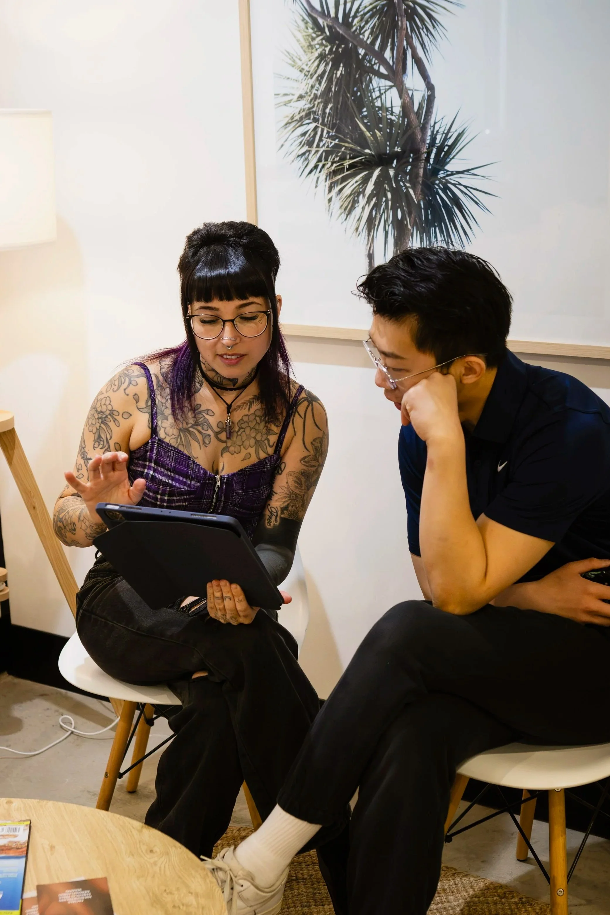 Two young adults sitting close together in a casual setting, engaged in a discussion while looking at a tablet. The woman has long dark hair with purple highlights, tattoos on her arms, and is wearing glasses and a purple checkered top. The man has short dark hair, glasses, and is wearing a dark shirt. There is a large artwork of a palm tree on the wall behind them, and they are seated on modern chairs around a small round table.