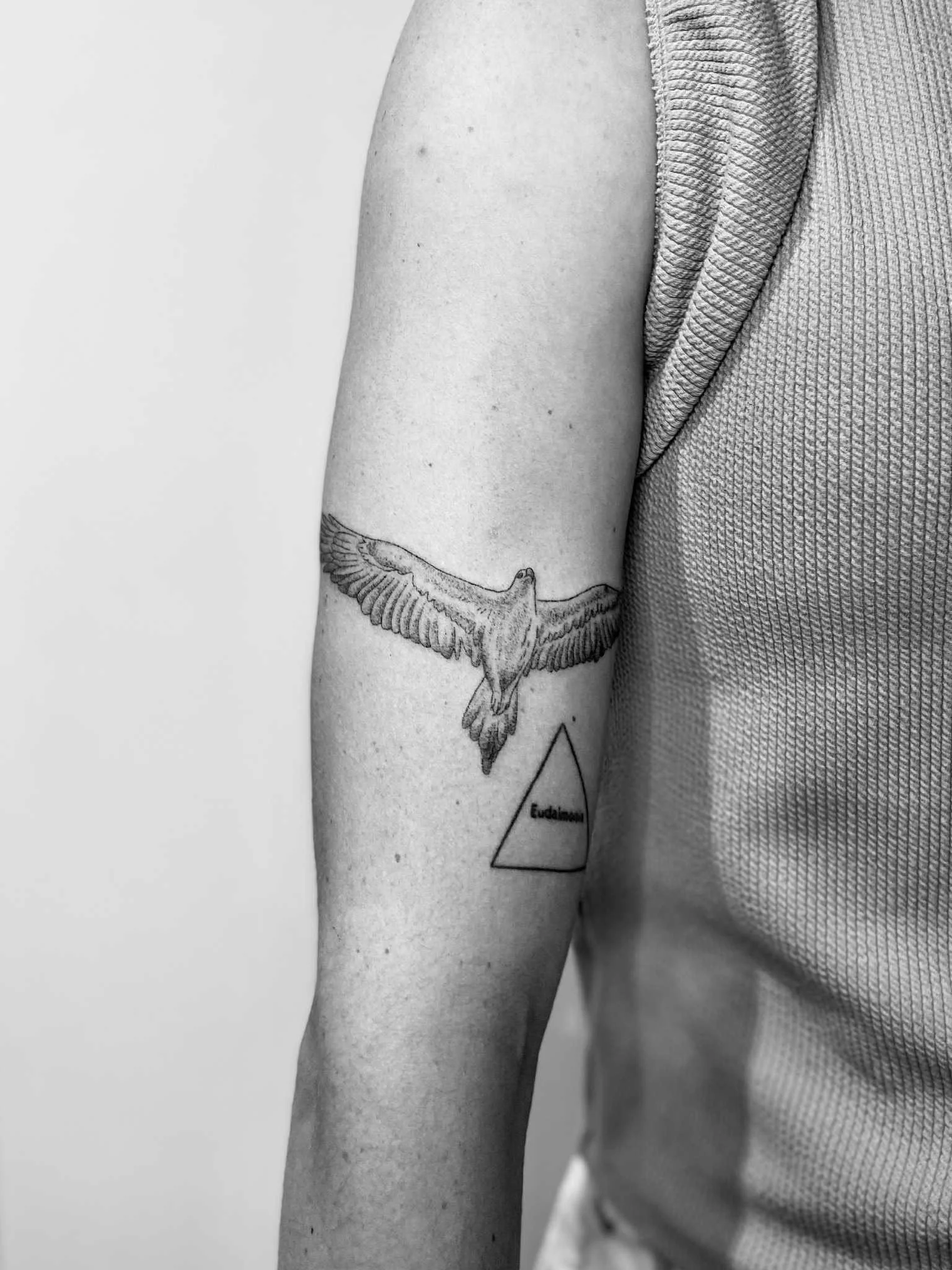 Black and white photograph of a person's bare arm displaying a tattoo of a bird in flight inside a triangle, with the label 'Eudoxum' beneath.