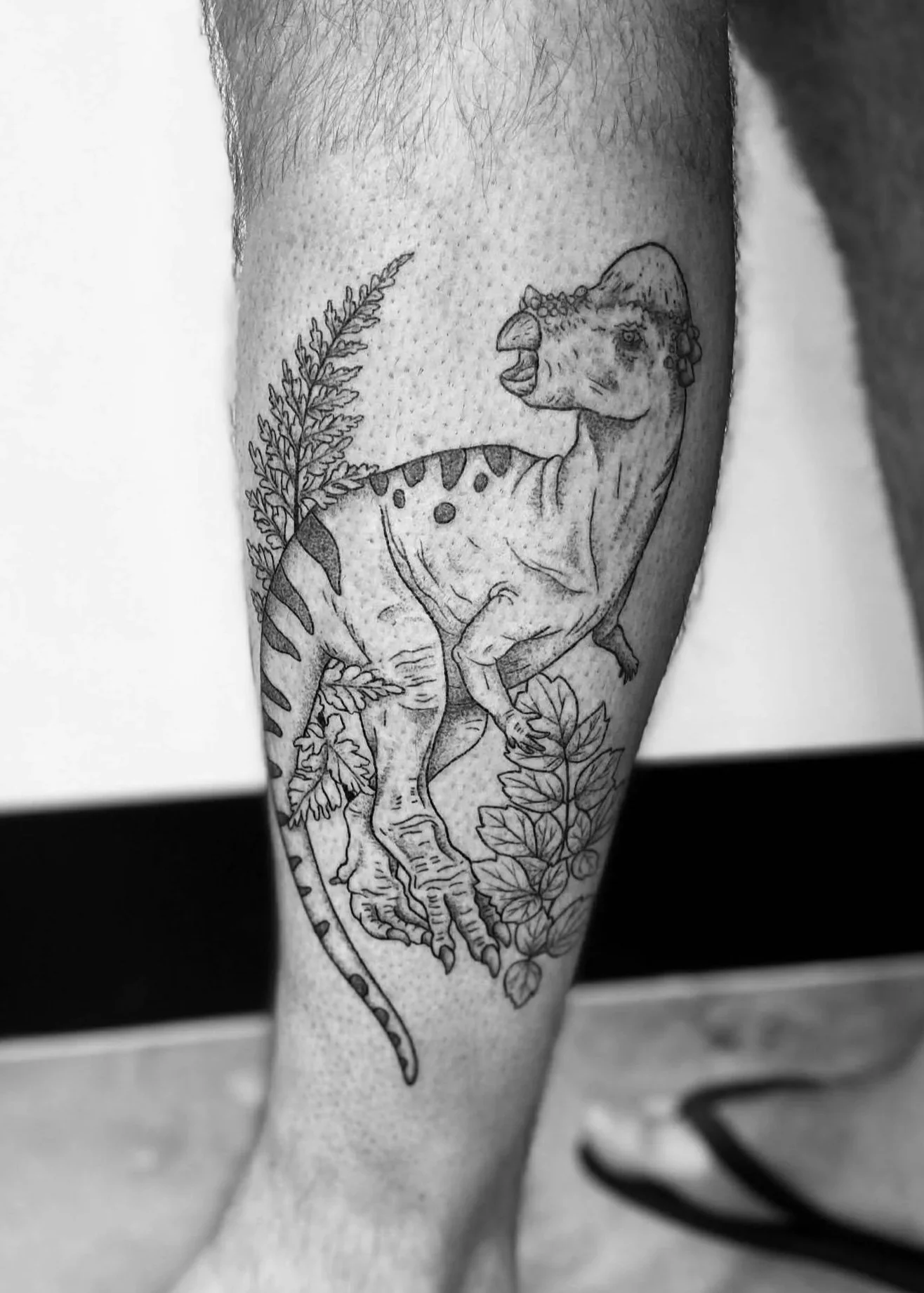 Black and white tattoo of a dinosaur, surrounded by plants.