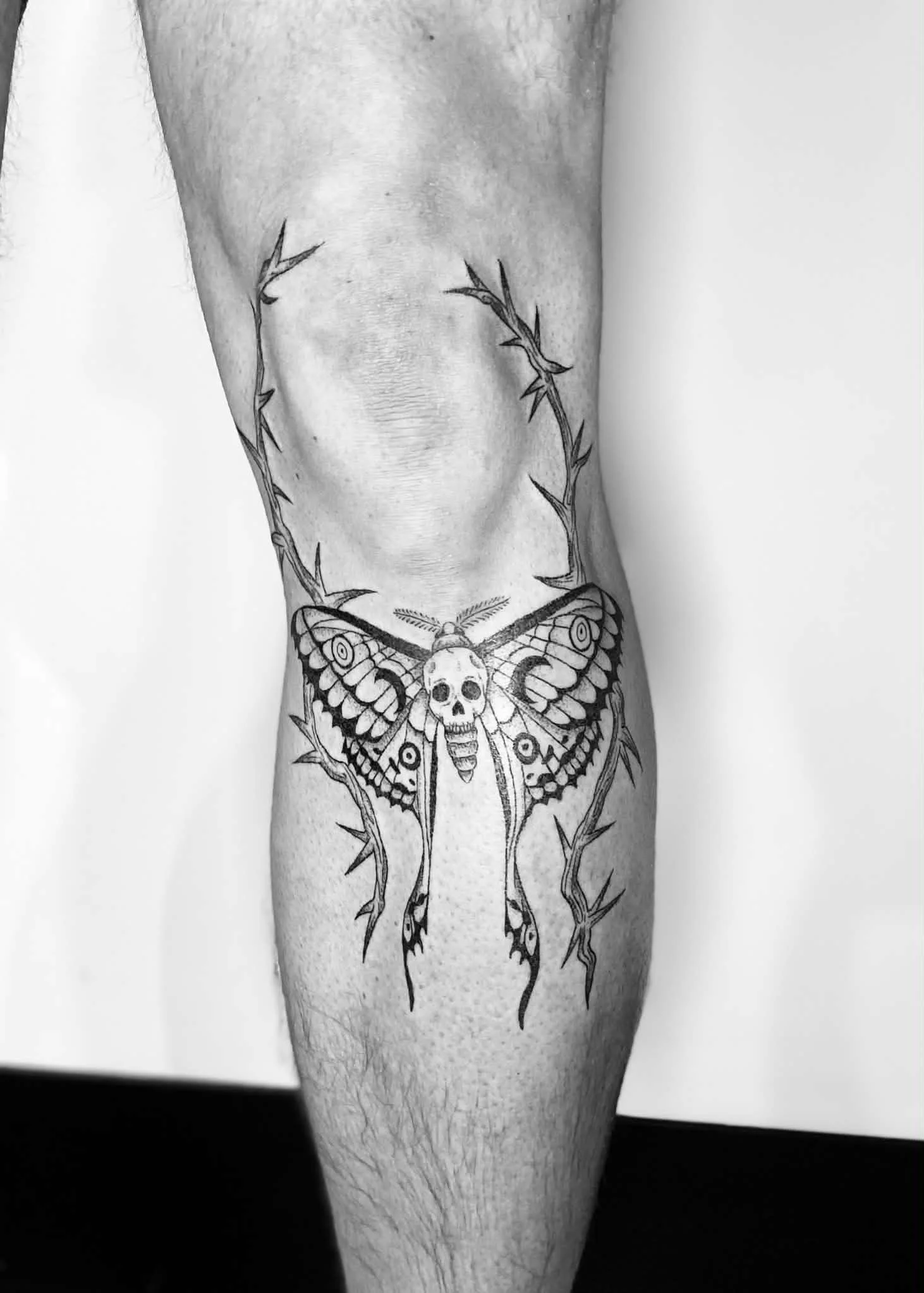 Black and white tattoo of a moth with a skull on its body, surrounded by thorns on a person's forearm.
