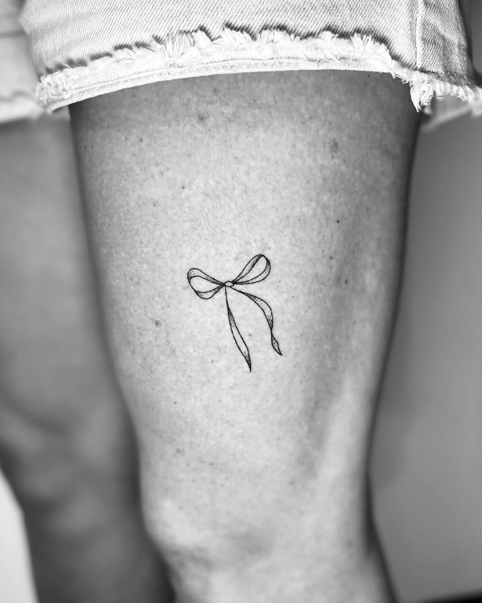 Close-up of a person's upper thigh and lower abdomen, with a tattoo of a bow on the skin. The person is wearing distressed denim shorts.