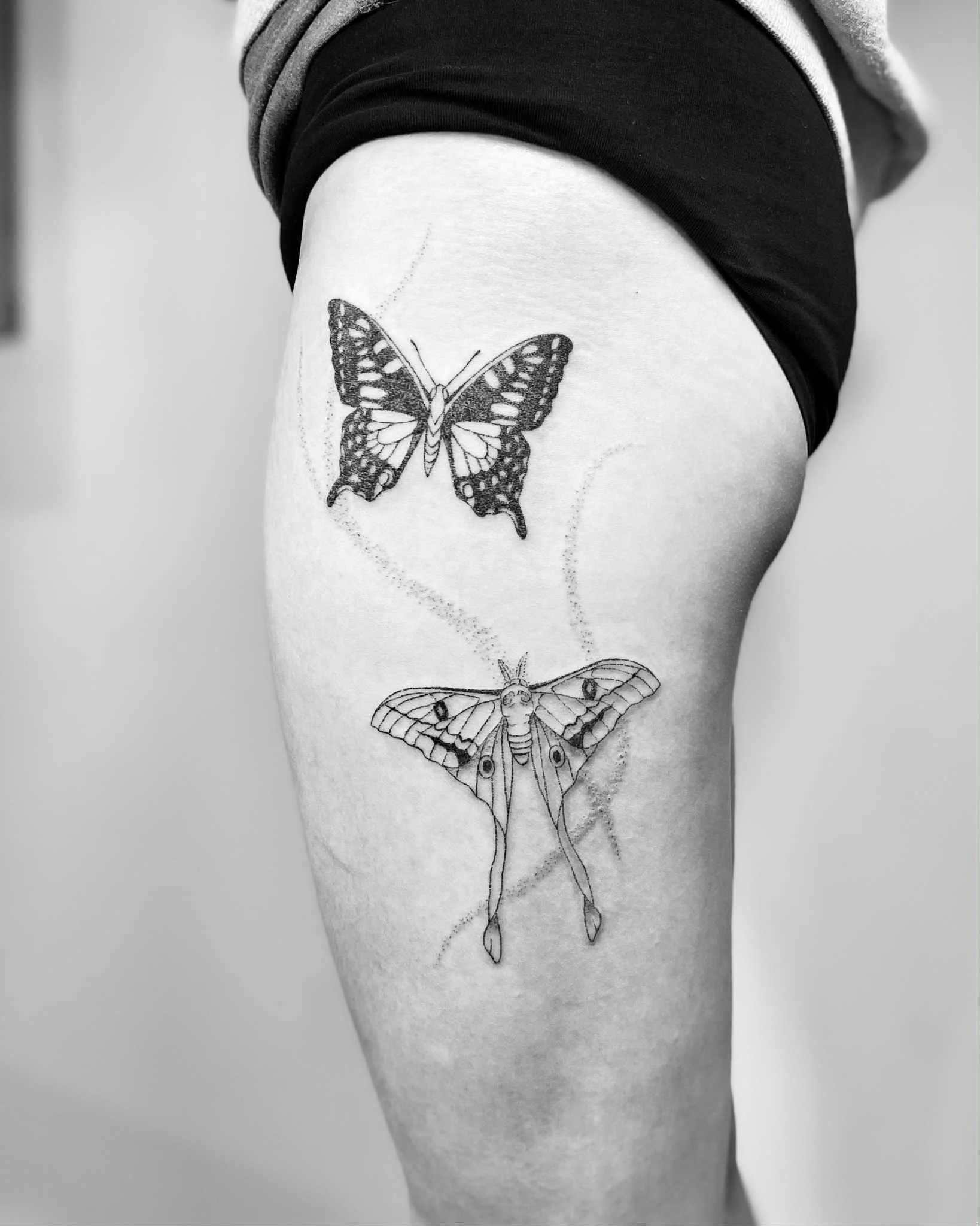 Black and white tattoo of a butterfly and a moth on a person's thigh, with the butterfly above the moth.