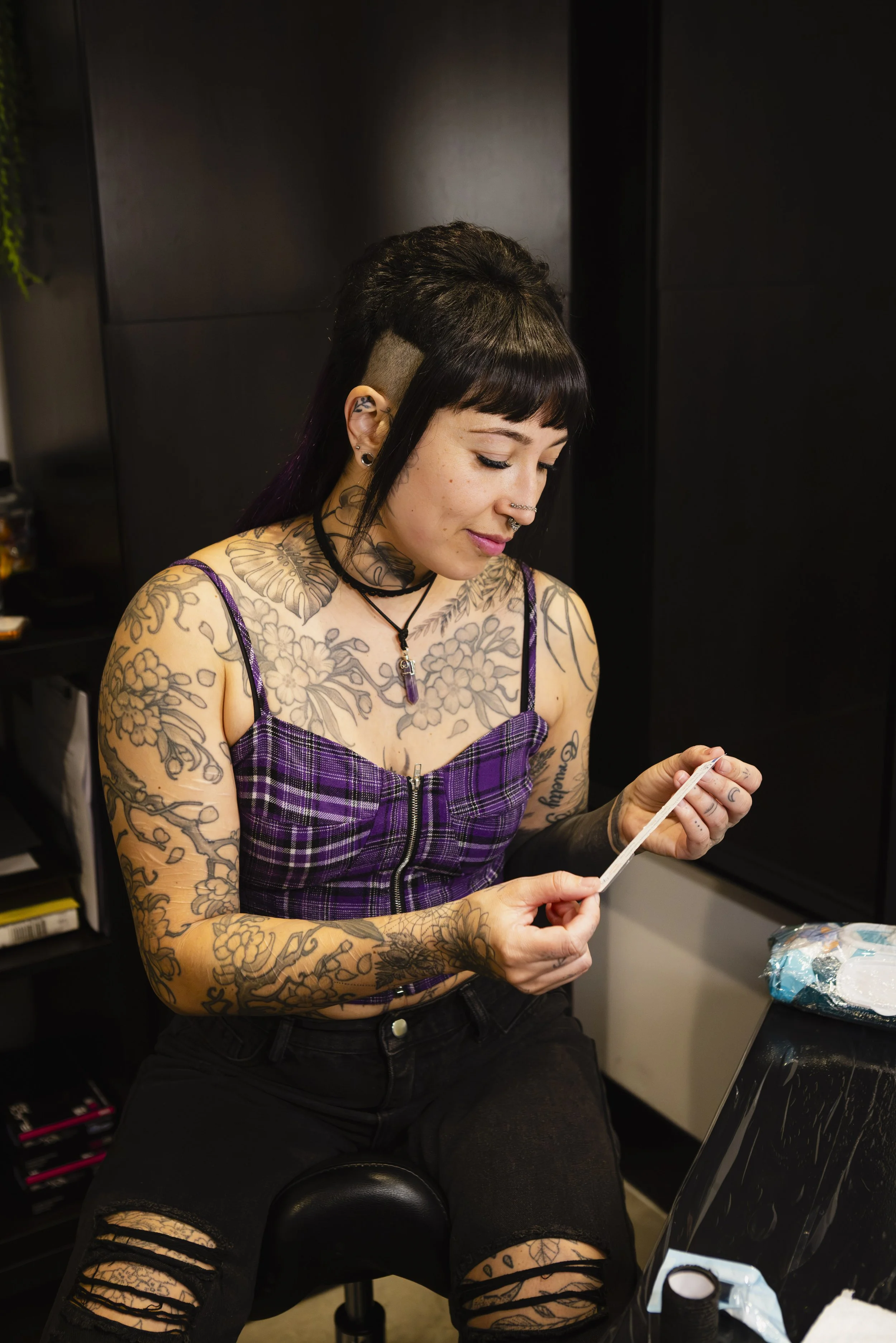A woman with tattoos, black hair with purple tips, and multiple piercings sits in a salon, looking down and holding a piece of paper.