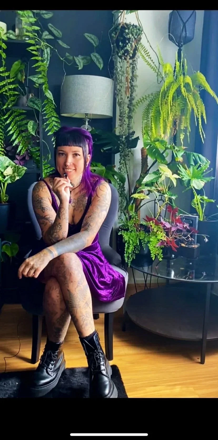 A woman with purple hair and tattoos sitting on a chair surrounded by green houseplants in a well-lit room.