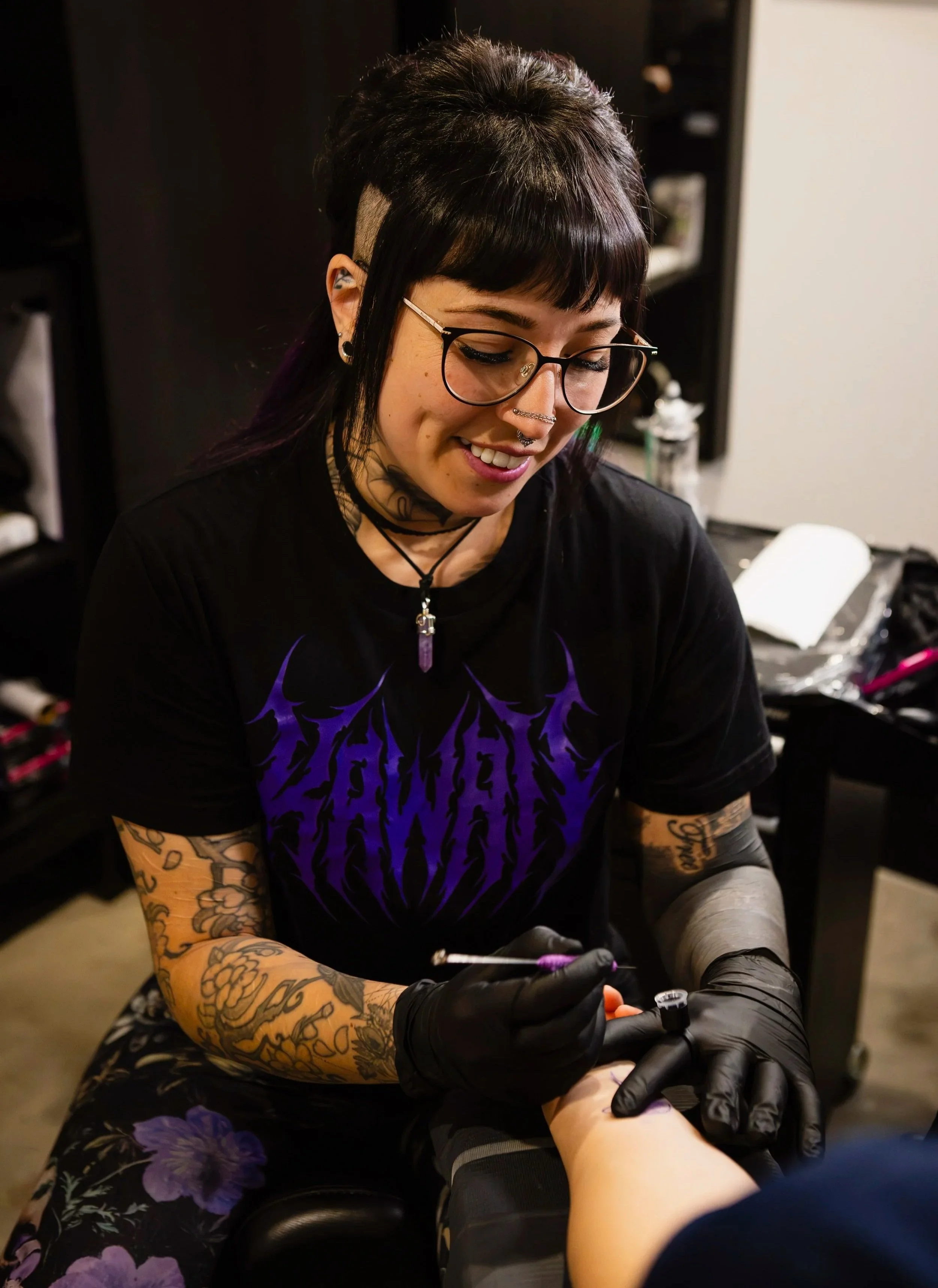 A woman with black hair, glasses, and tattoos getting a tattoo on her forearm done by an artist in black gloves at a tattoo studio.