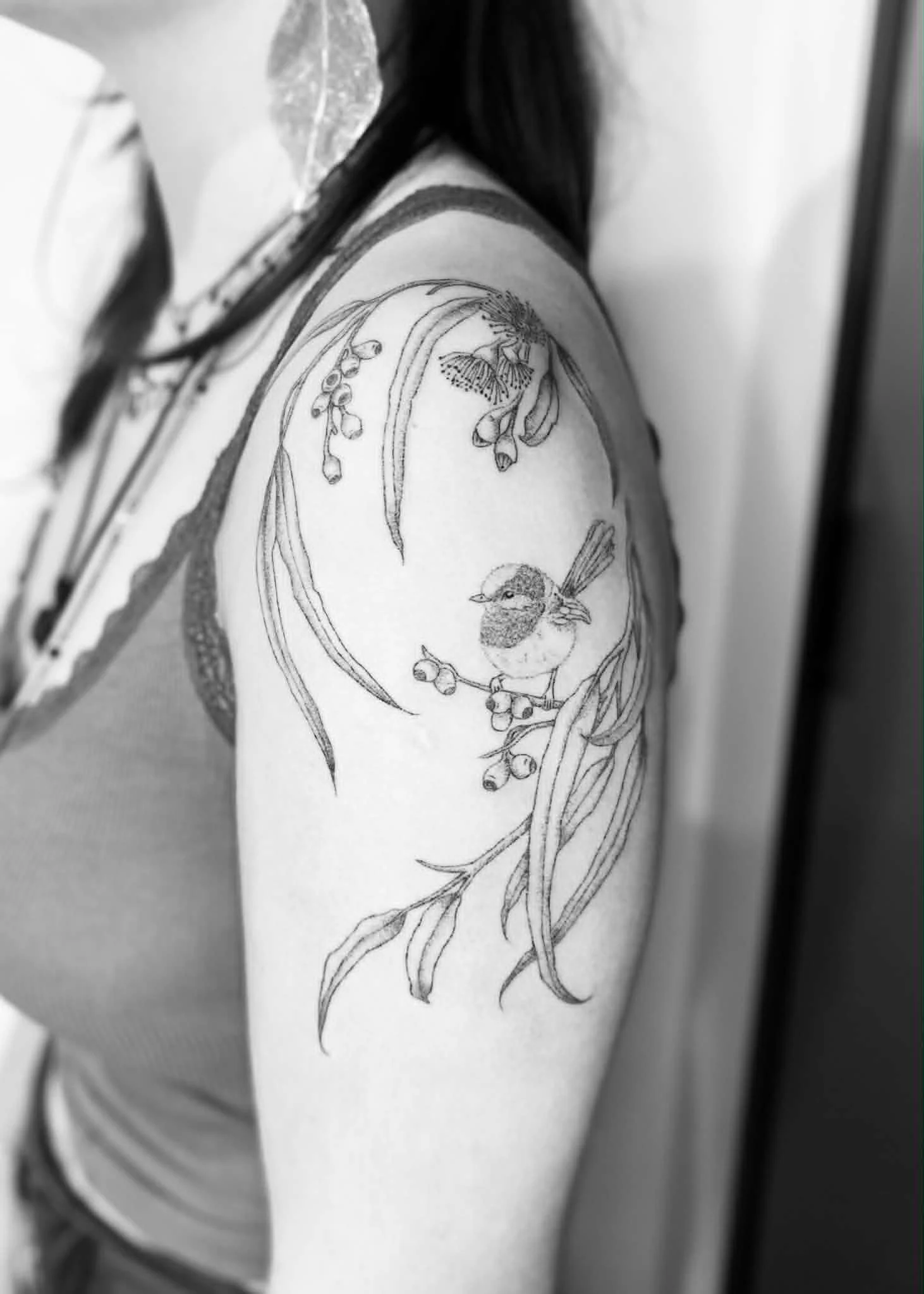 Line art tattoo on person's shoulder of a small bird perched on a branch with leaves and berries, with a larger bird flying above.