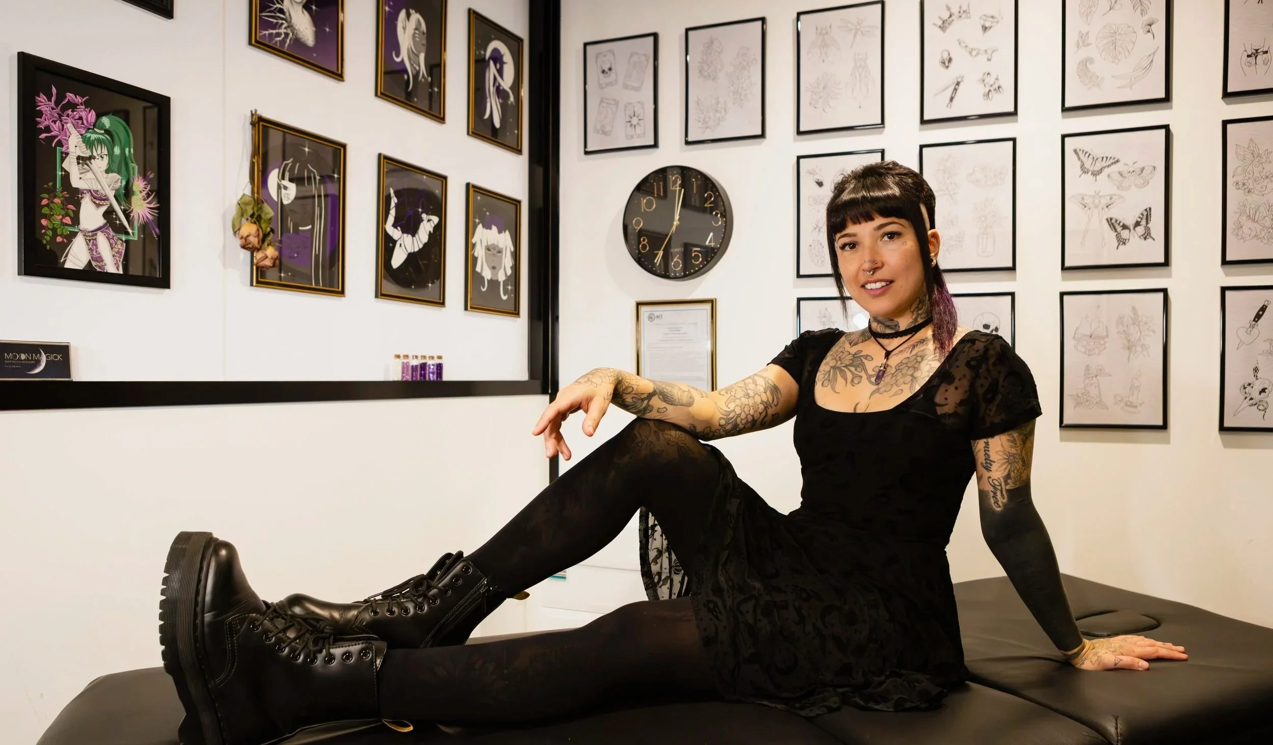 Young woman with short dark hair, tattoos, wearing black lace dress and combat boots, sitting on a black table in an art gallery with framed artwork and a clock on the wall behind her.