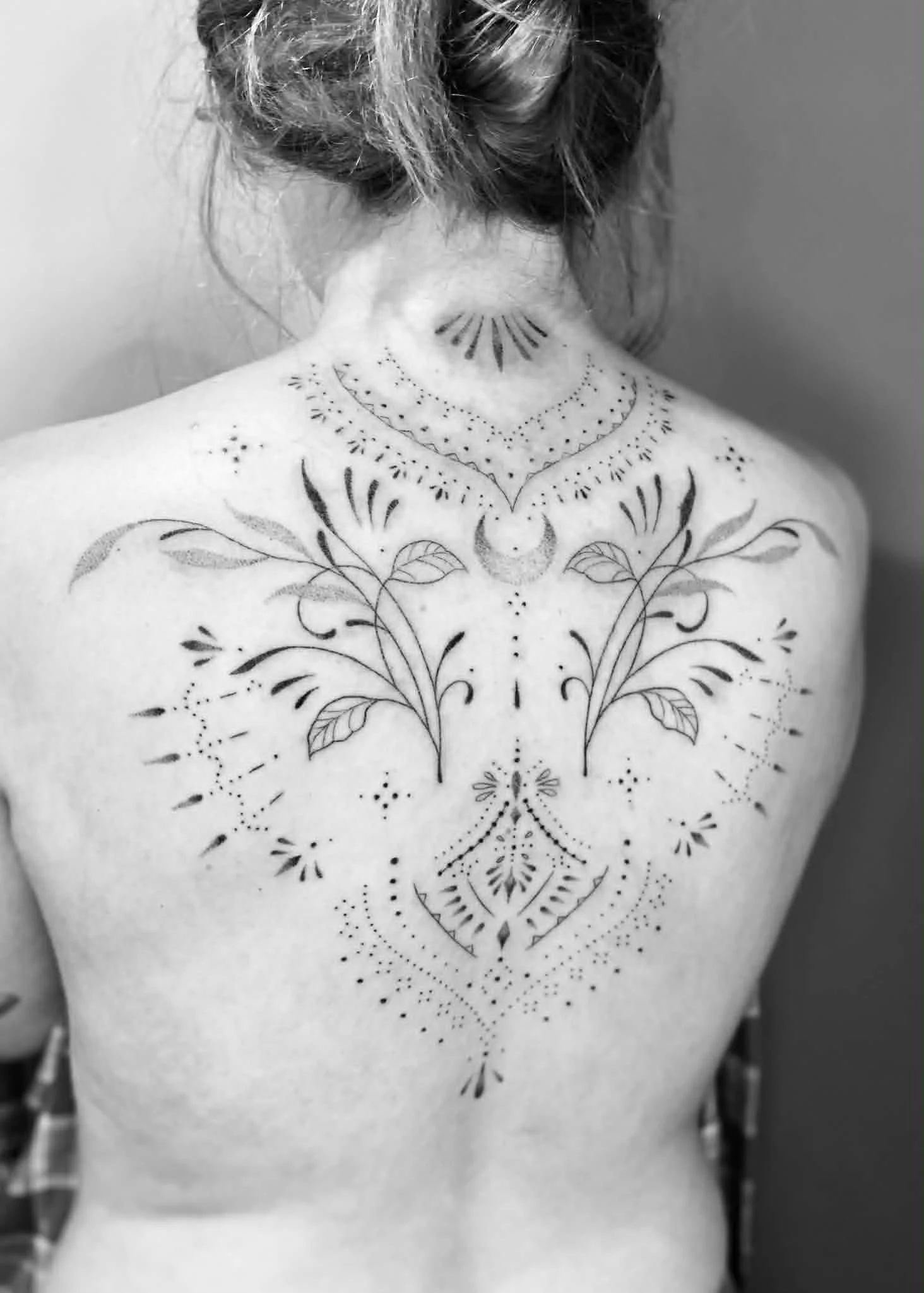 A person with a hairstyle tied up, showing their back and shoulders with a temporary tattoo of a symmetrical, floral and geometric design on their upper back and neck in black ink.