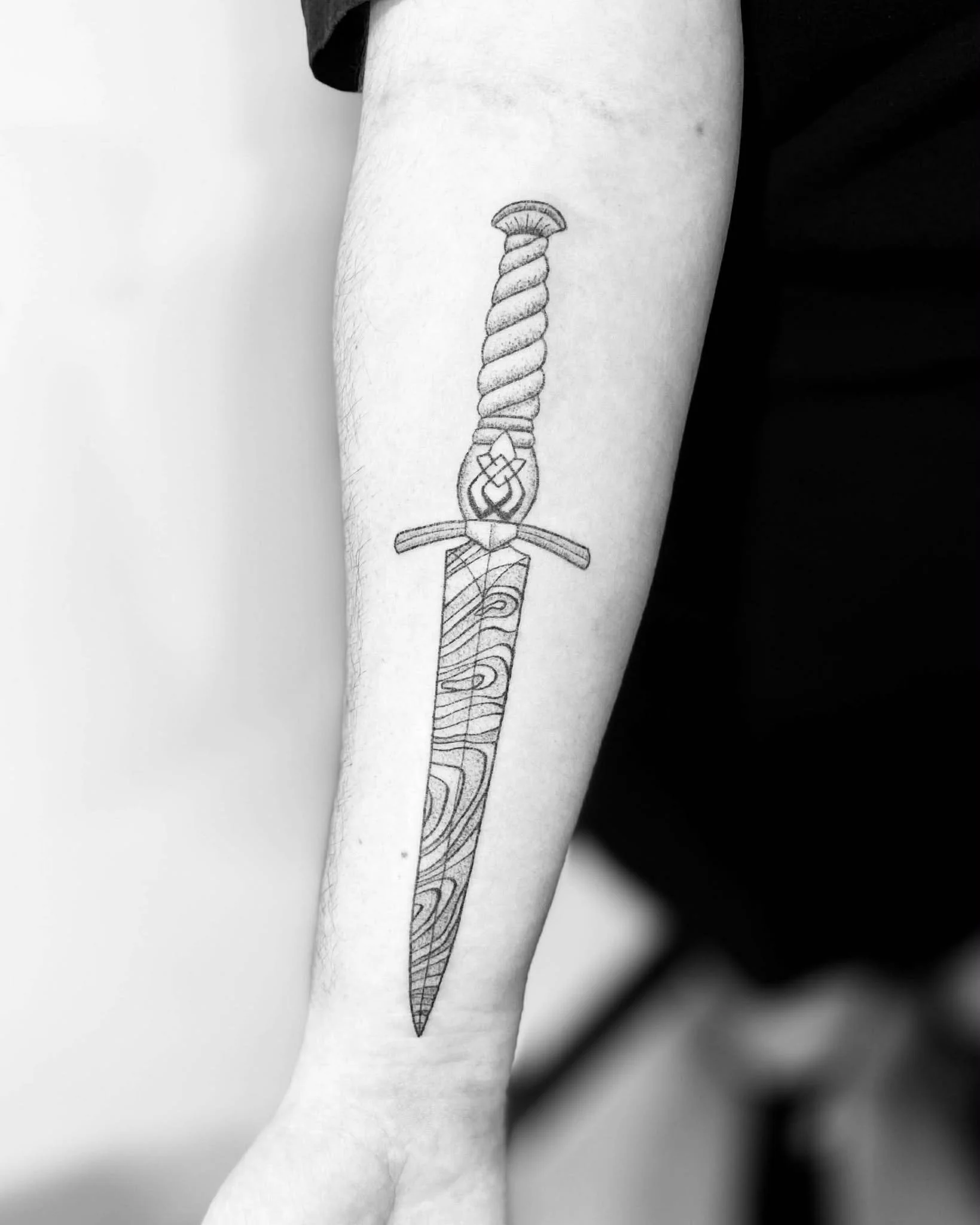 Black and white photo of a forearm with a tattoo of a sword, featuring a detailed hilt and a wood grain pattern on the blade.