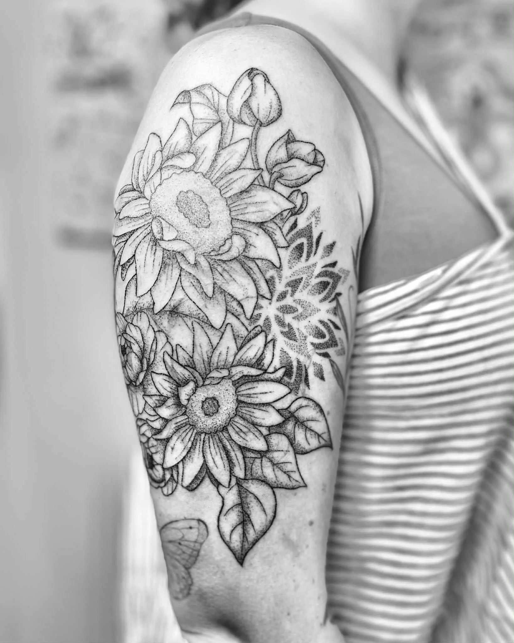 Black and white photo of a person's upper arm tattoo featuring detailed sunflowers, buds, and leaves.