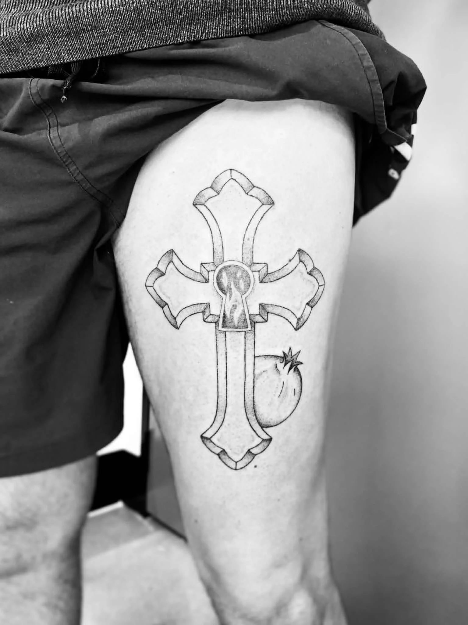 A black and white photo of a person's thigh, showing a tattoo of a cross with a circular element in the center and a small flame at the bottom.