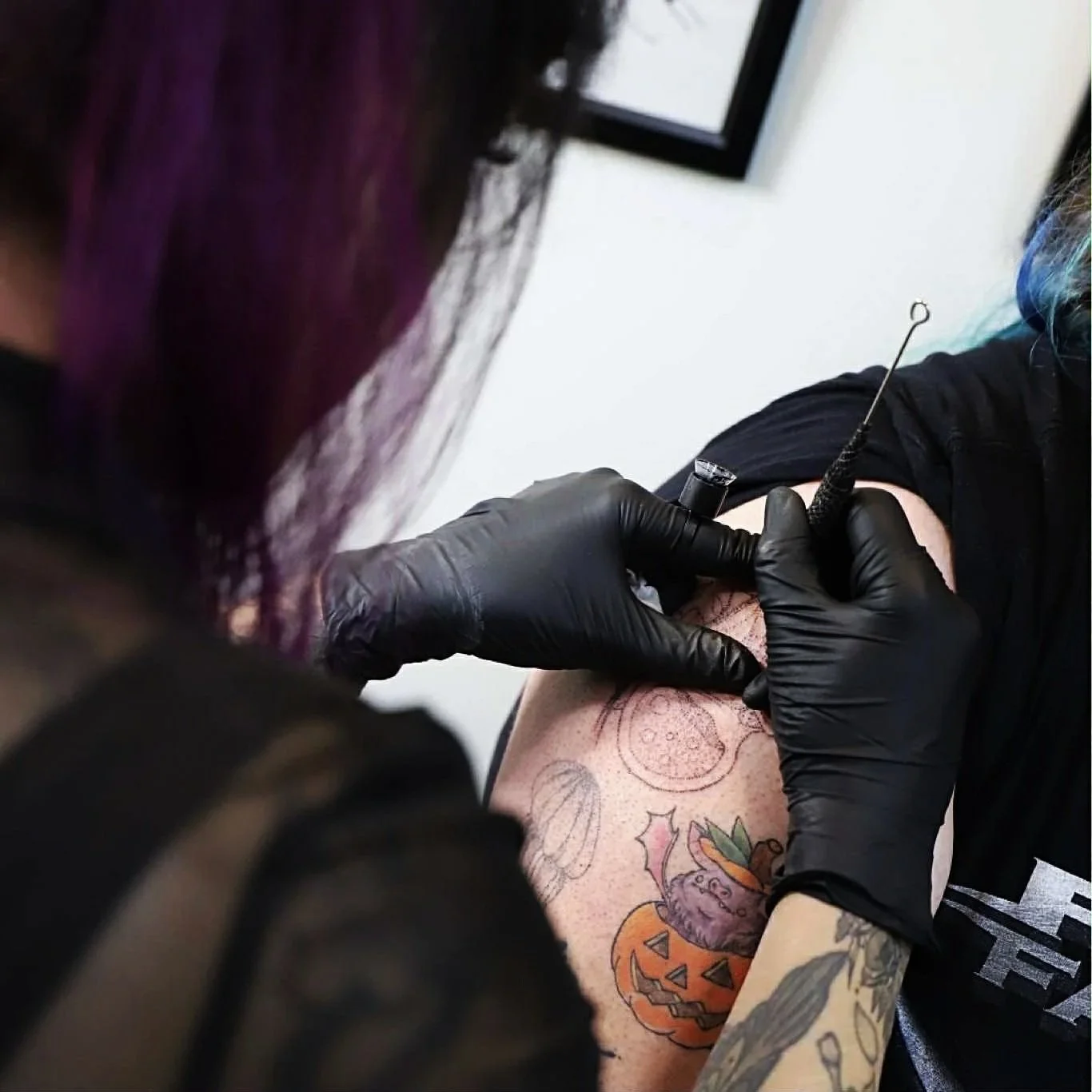 A person with colorful hair getting a tattoo on their arm by a tattoo artist wearing black gloves.
