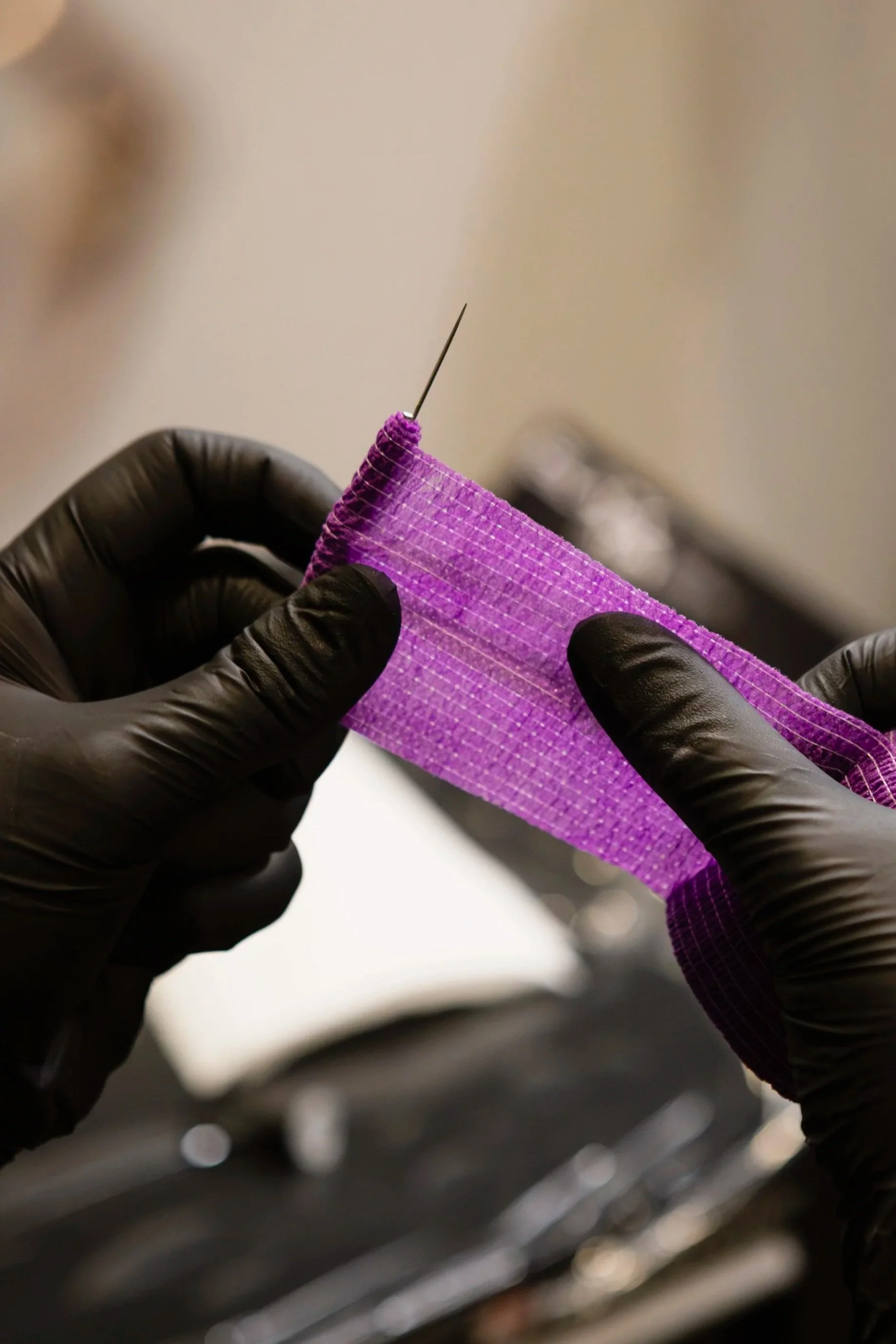 Close-up of hands wearing black gloves holding a purple textured surface with a sewing needle inserted into it.