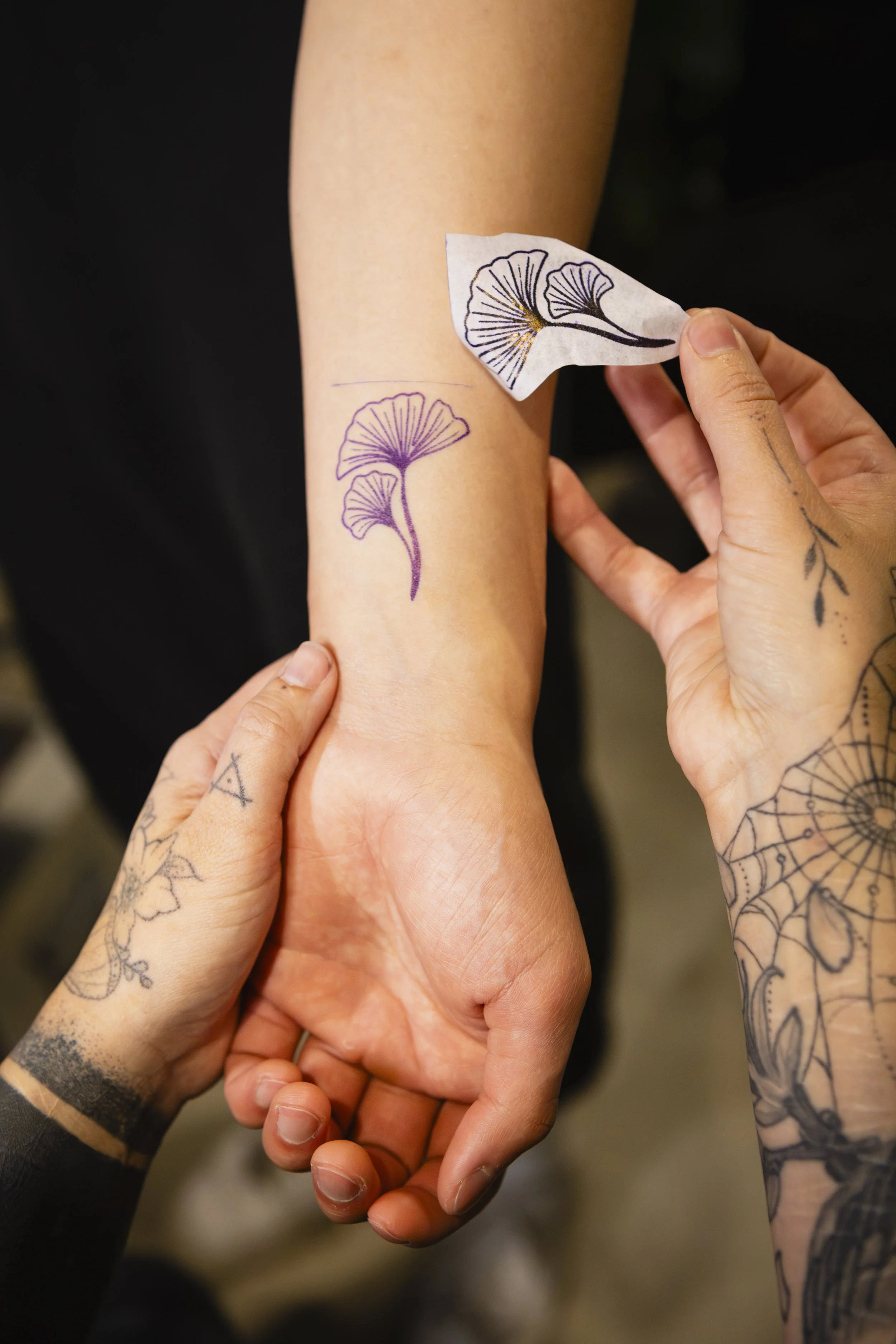 Person applying temporary tattoo of a ginkgo leaf to another person's forearm, with tattoos visible on their hands and arms.