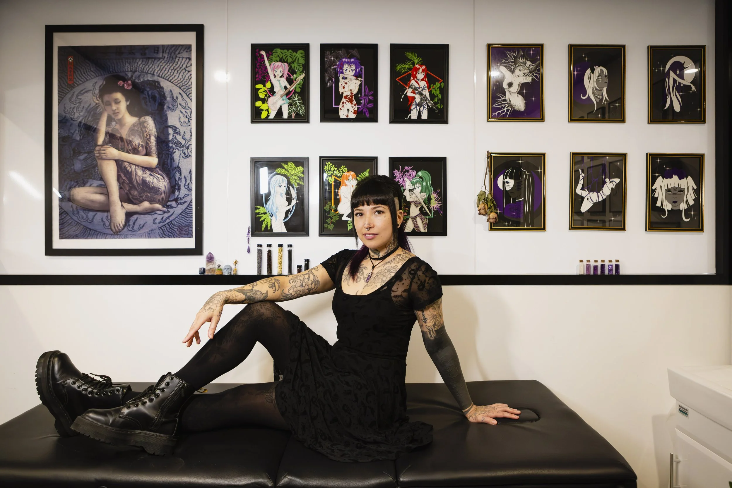 A woman with black hair and tattoos on her arms and neck, dressed in black lace clothing and black combat boots, seated on a black padded bench in front of a white wall with framed artwork. The artwork features various colorful illustrations of anime-style characters and a large portrait of a woman with dark hair and traditional clothing.