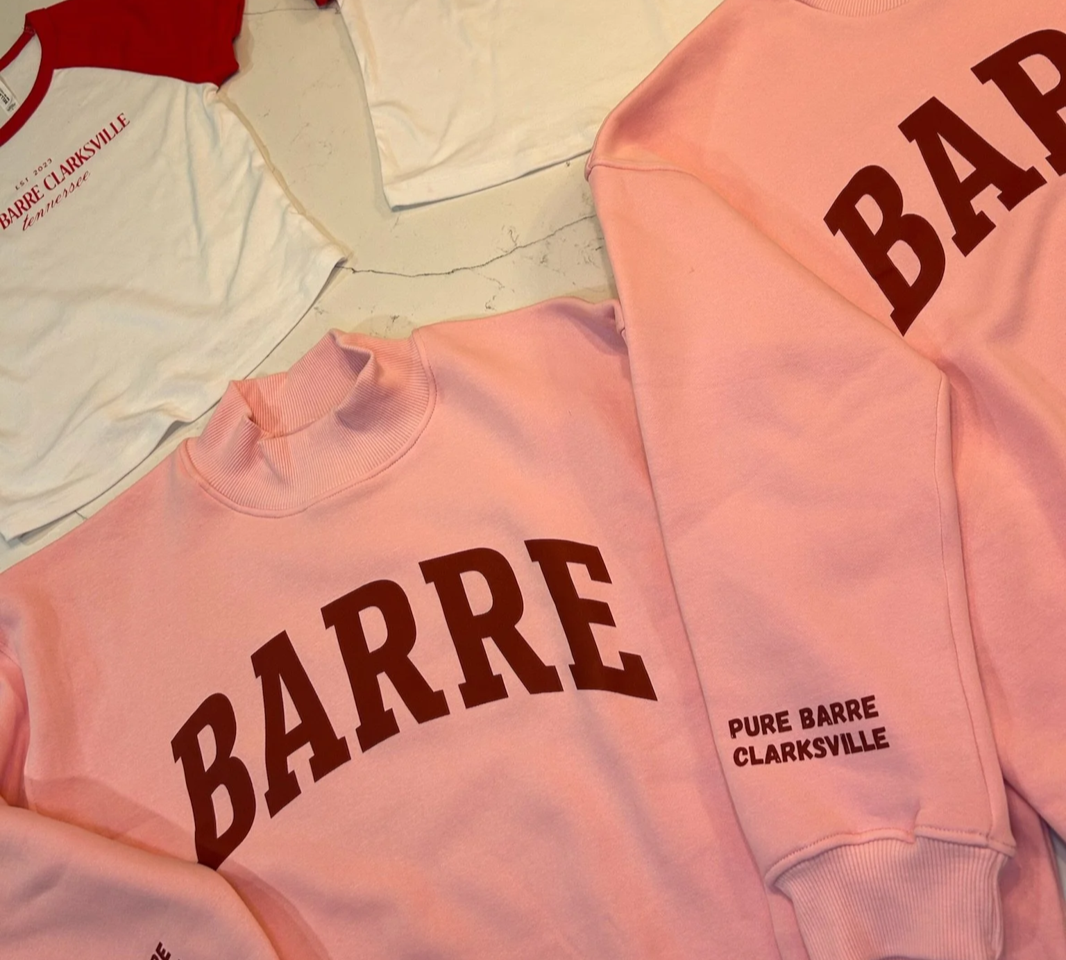 Pink sweatshirt with large dark red text 'BARRE' and smaller black text 'PURE BARRE CLARKSVILLE,' alongside a white t-shirt with red text 'PURE BARRE CLARKSVILLE' and red trim on the collar, laid out on a surface.
