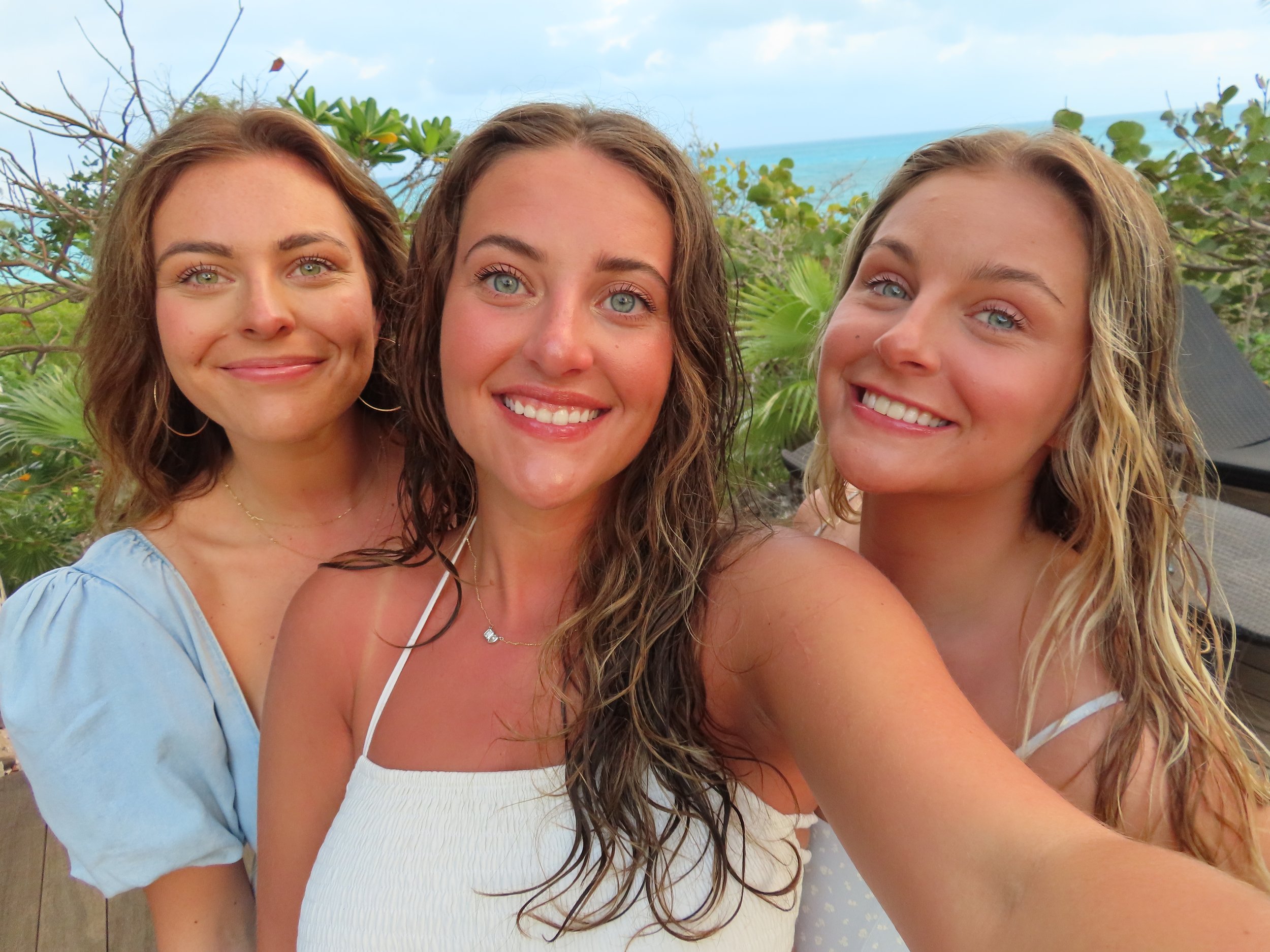 Three women taking a selfie outdoors near the ocean, with greenery and trees in the background, smiling and enjoying a sunny day.