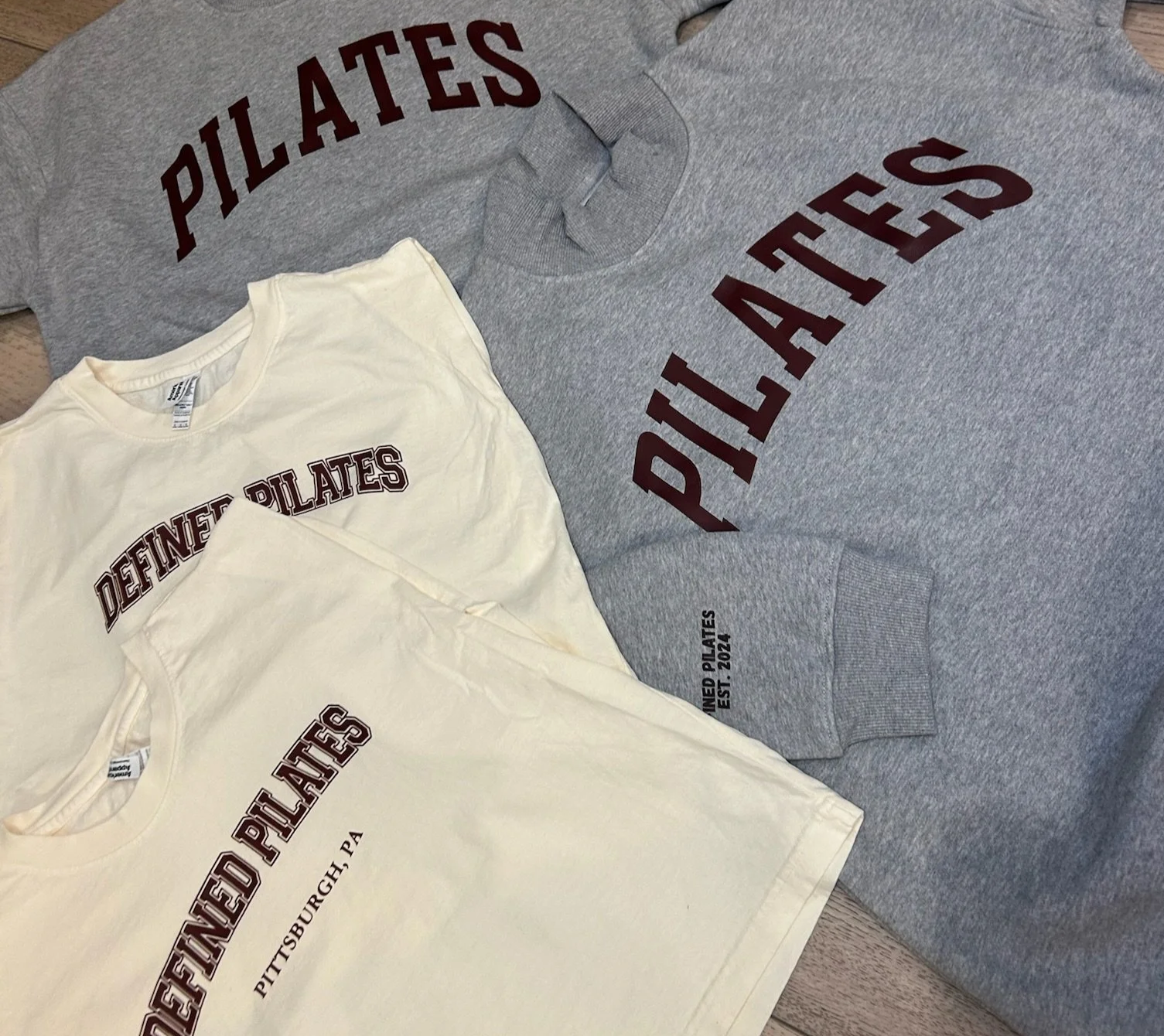 Gray and white t-shirts with 'DEFINED PILATES' in maroon text, laid out on a wooden surface.