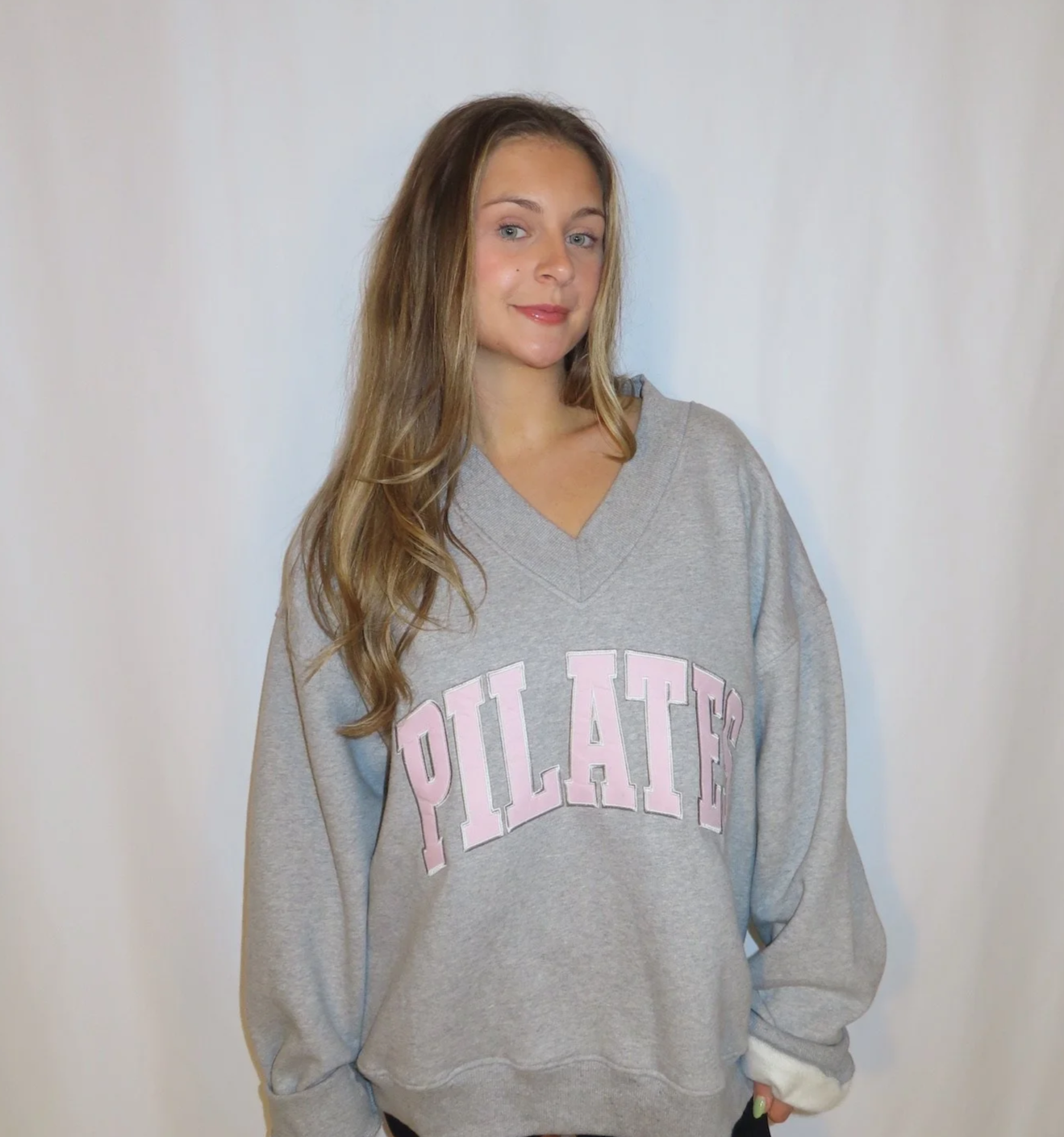 A young woman with long, wavy brown hair standing in front of a plain light-colored wall, wearing a gray oversized sweatshirt with the word 'PLATES' in pink lettering.