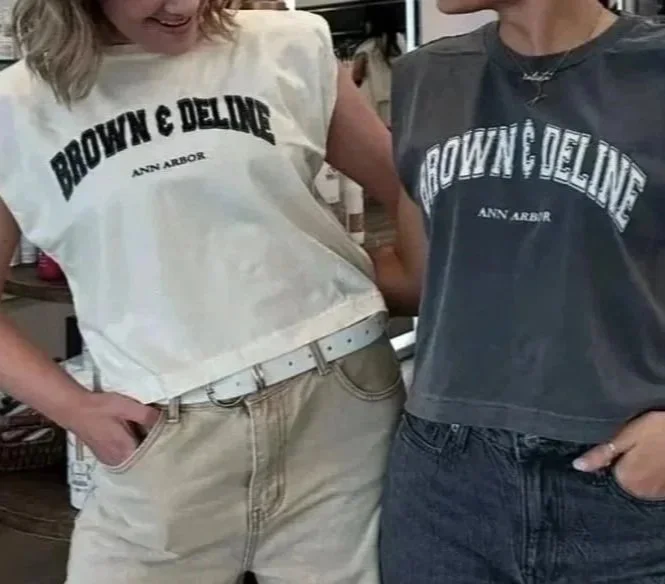 Two people are wearing matching T-shirts with the words 'BROWN & DELINE' and 'ANN ARBOR' printed on them. One is wearing a white shirt, and the other is wearing a dark shirt. They have their hands in their pockets and are standing close together.