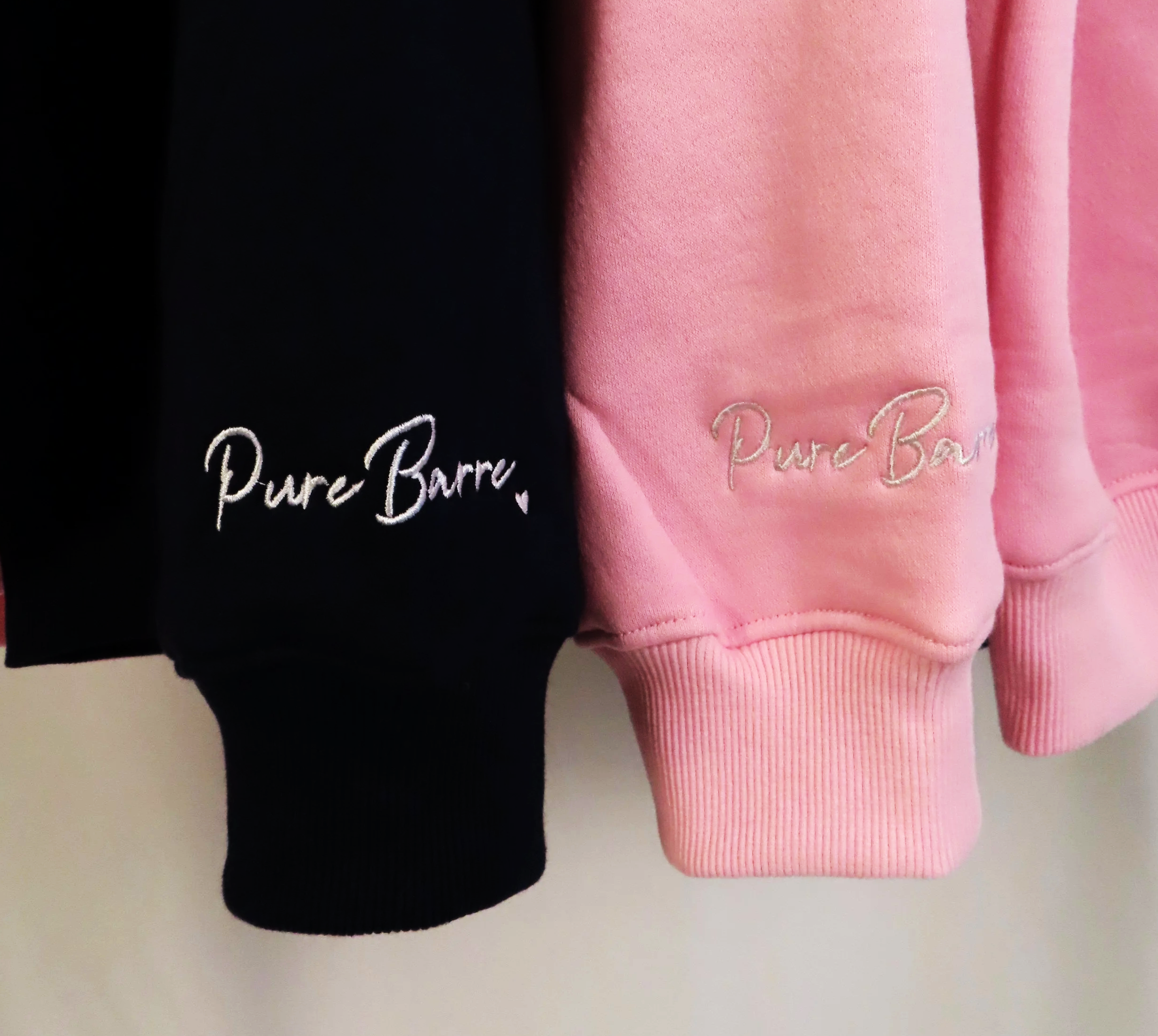 Close-up of two sweatshirts, one black and one pink, with embroidered text 'Pure Barre' on the lower part of each sleeve.
