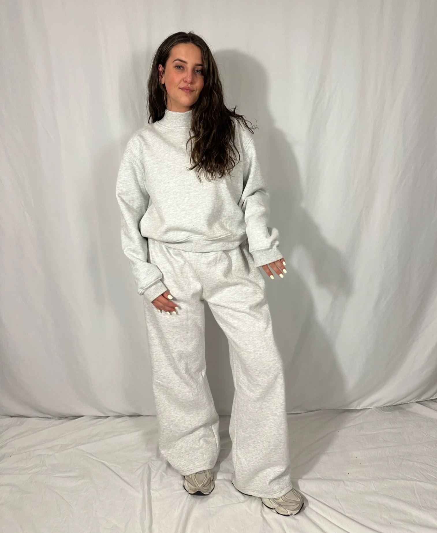 A woman with long wavy hair wearing a white, oversized sweatshirt and matching sweatpants, standing against a plain white backdrop.