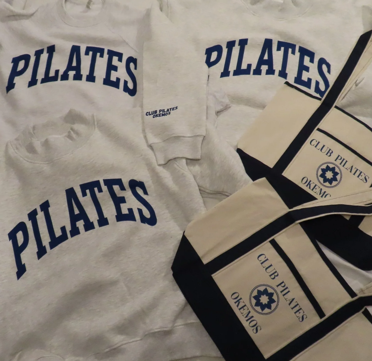 White clothing and cloth items with the words 'PILATES' and 'CLUB PILATES OKEMO' printed in blue, and a logo featuring a blue and white star-like design.