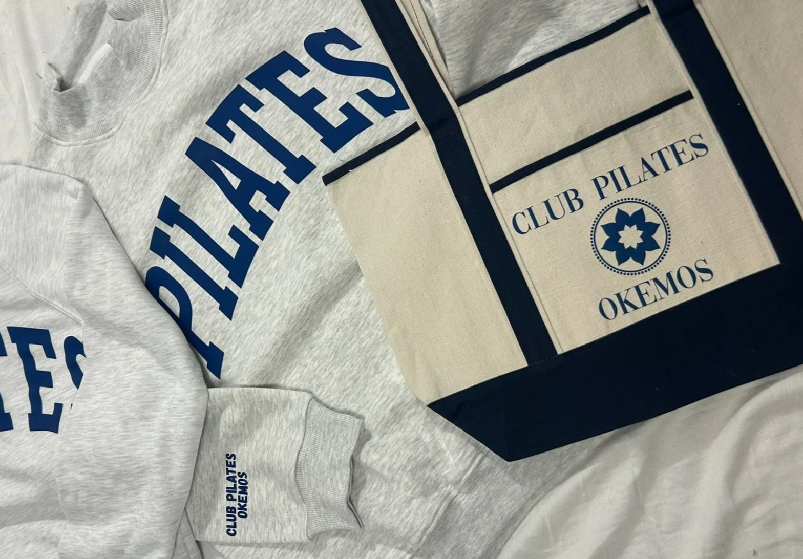 Gray sweatshirt with blue text 'PALLAS', beige tote bag with blue text 'CLUB PILATES OKEMOS' and a blue decorative emblem.