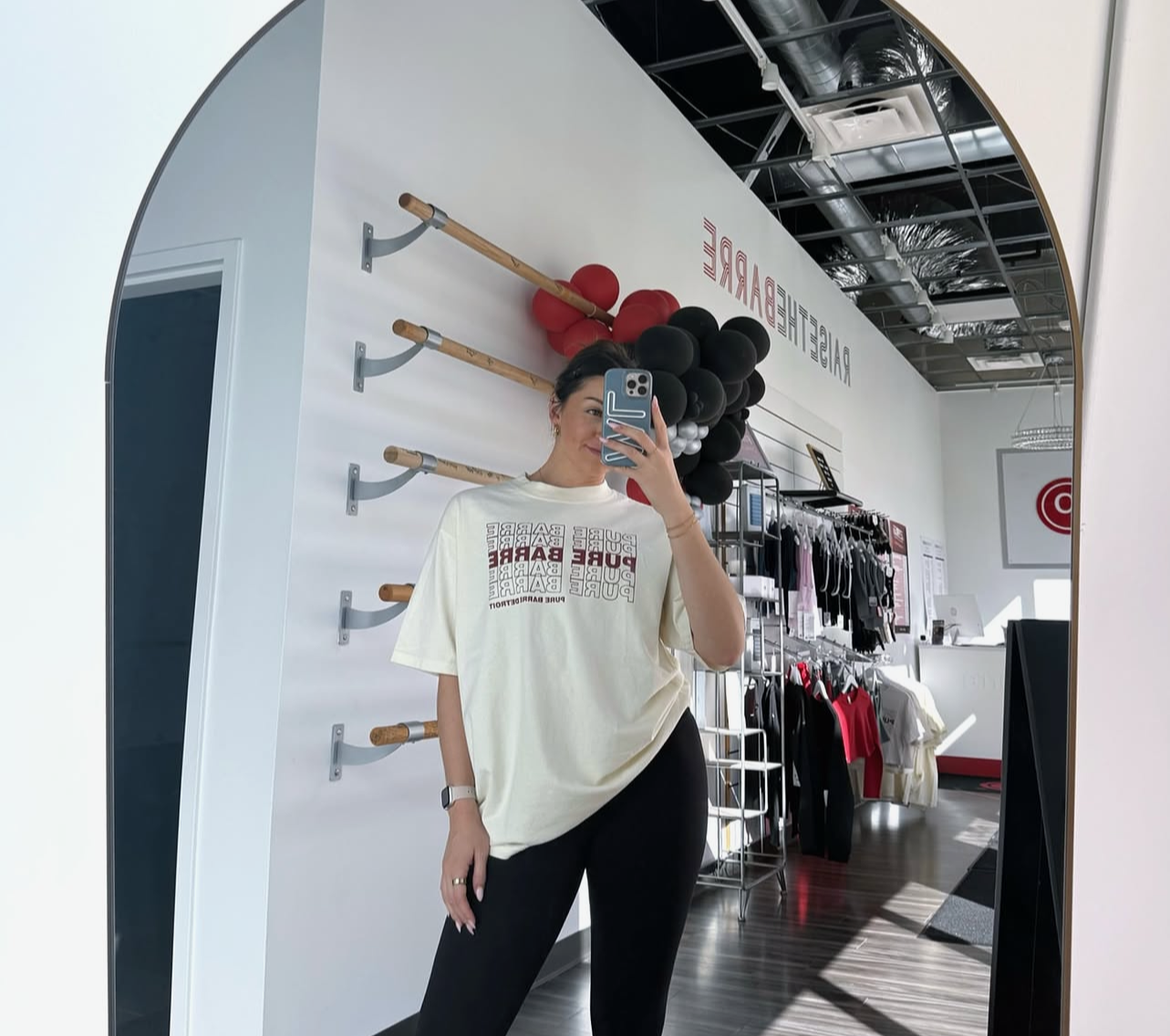 A woman taking a mirror selfie inside a clothing store, wearing a white graphic t-shirt and black pants, with gym equipment and clothing racks visible in the background.