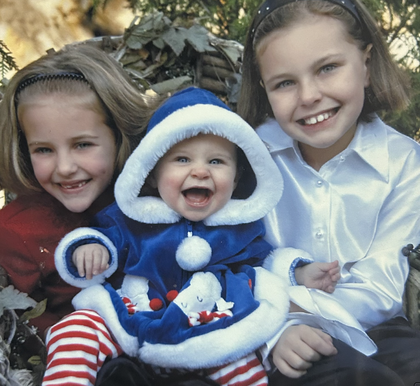 Three children smiling, sitting outdoors among fallen leaves, with trees in the background. The youngest in the middle is wearing a blue and white winter coat with a hood, with snowman design, and red and white striped pants. The girl on the right is wearing a white satin shirt, and the girl on the left is wearing a red jacket.