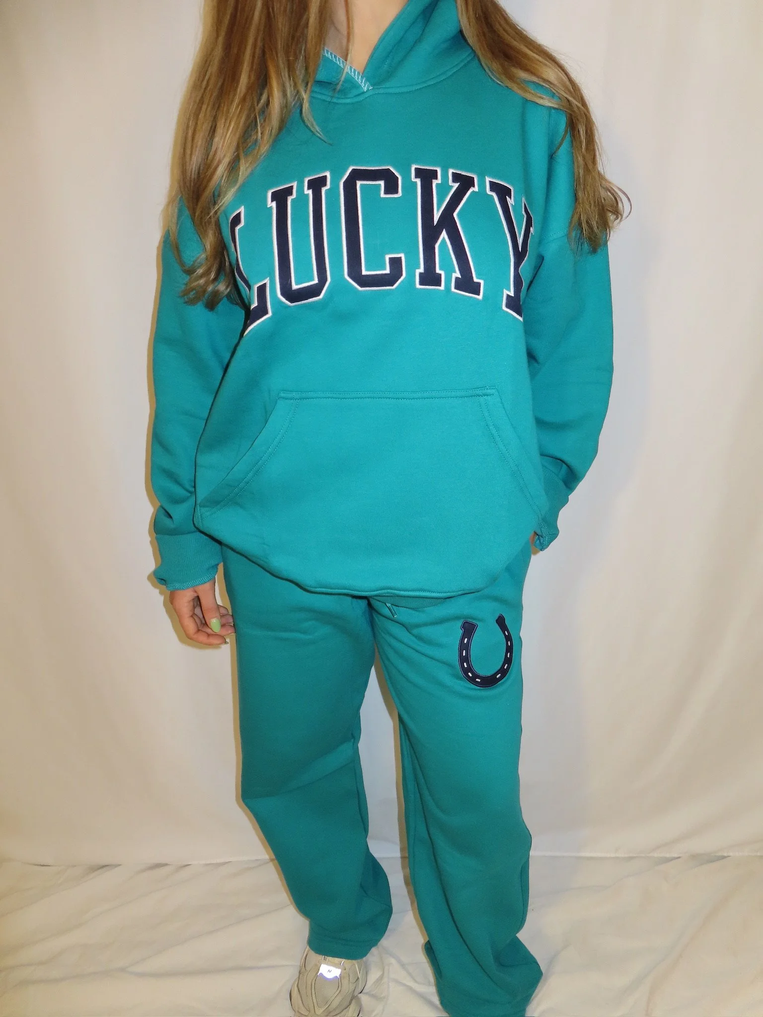 Person wearing a teal hoodie with the word 'LUCKY' on the front and teal sweatpants with a horseshoe design, standing indoors against a plain wall.