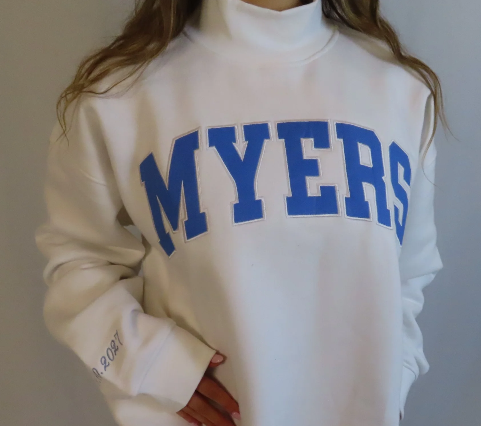 A person wearing a white sweatshirt with large blue letters spelling 'MY HERO' on the front. The person's left arm is bent with the hand resting near the waist.