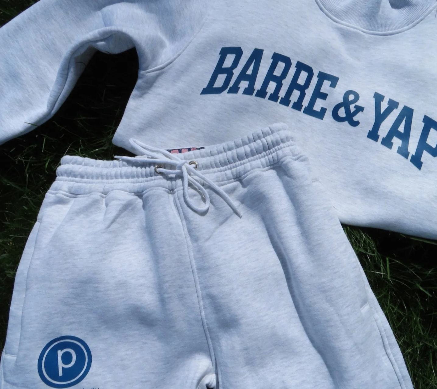 A light gray sweatshirt and matching sweatpants laid out on grass. The sweatshirt has blue lettering that partially reads 'BARRIE&YARD.' The sweatpants have a blue circular logo with a lowercase letter 'p' inside.