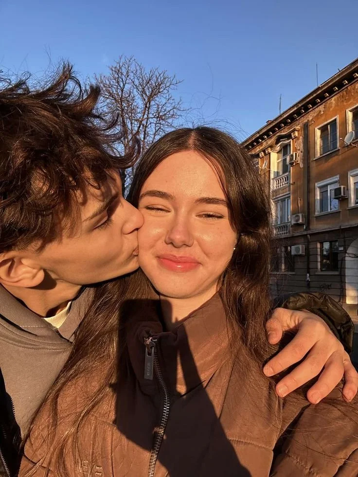 A young man giving a kiss on the cheek to a young woman with long brown hair, both smiling, outdoors during sunset. The woman is wearing a brown jacket and the man is wearing a hoodie. In the background, there are buildings and a clear blue sky.