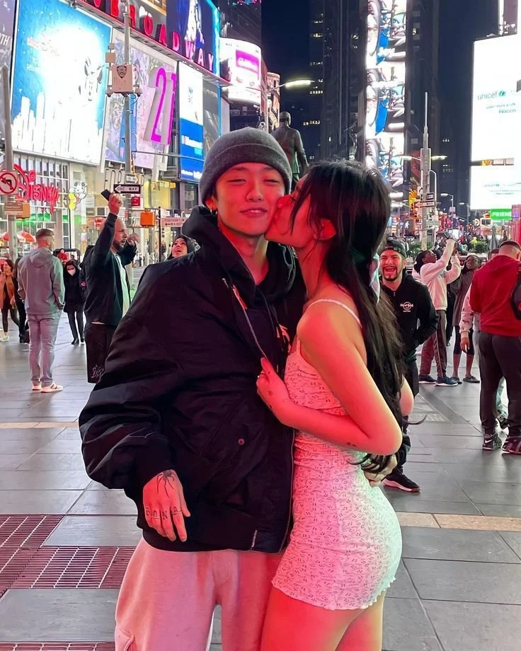 A young woman is kissing a young man on the cheek in a busy city street at night, illuminated by bright neon lights and digital billboards. The man is smiling with closed eyes, wearing a gray beanie and a black jacket. The woman is in a light-colored, textured dress, with her arms around the man's waist. There are several other people in the background, some taking photos and others walking, with tall buildings and glowing signs characteristic of Times Square in New York City.
