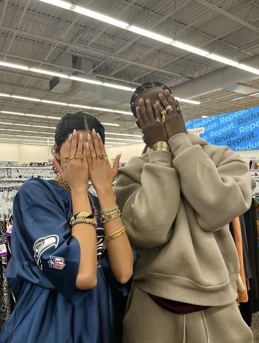Two people standing in a retail store, covering their faces with their hands, wearing multiple rings and bracelets, with clothing racks and a blue Reuse sign in the background.