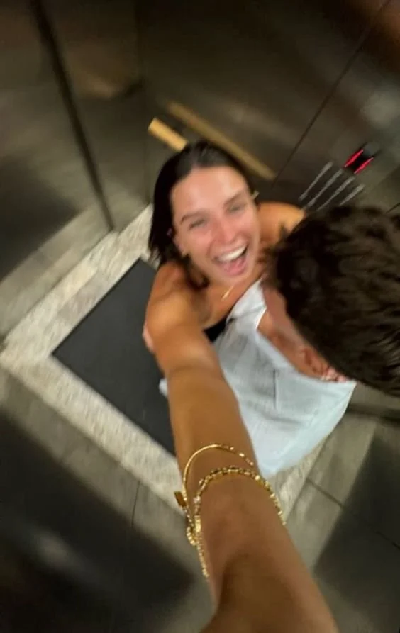 A woman laughing and smiling, being lifted or held by a person whose arm is visible, in an elevator with dark wood walls and a black mat on the floor.