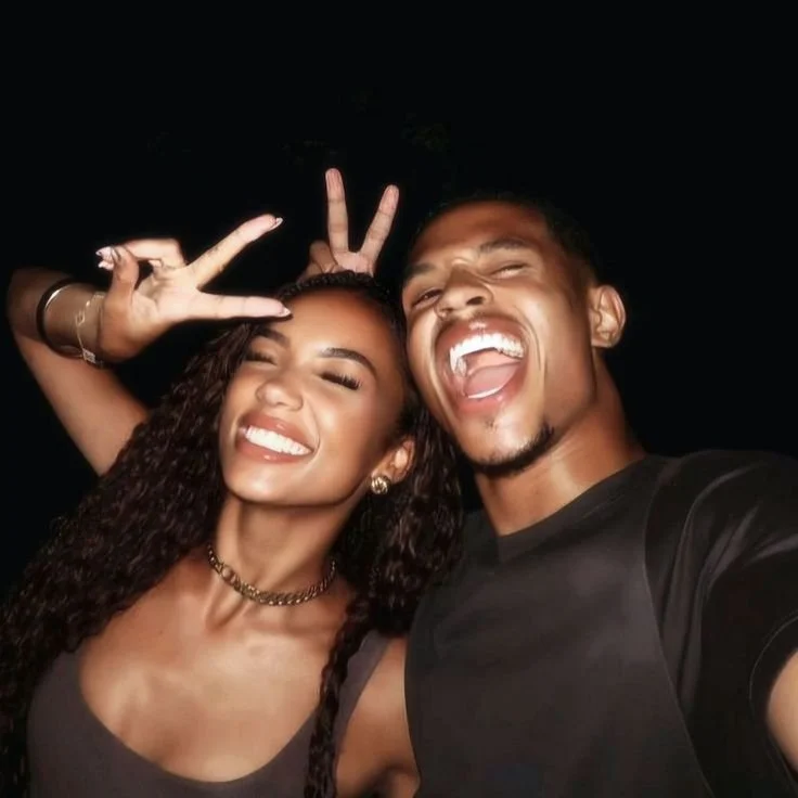 A young man and woman smiling and making playful gestures in a selfie, with a dark background.