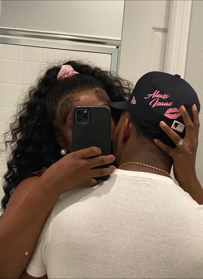 A woman taking a mirror selfie with a man, both holding each other close. The woman has curly hair in a ponytail with a pink scrunchie and is wearing large pearl earrings. The man is wearing a black baseball cap with pink embroidery that says 'Alexis Isaac' and kiss lips, and a white T-shirt.